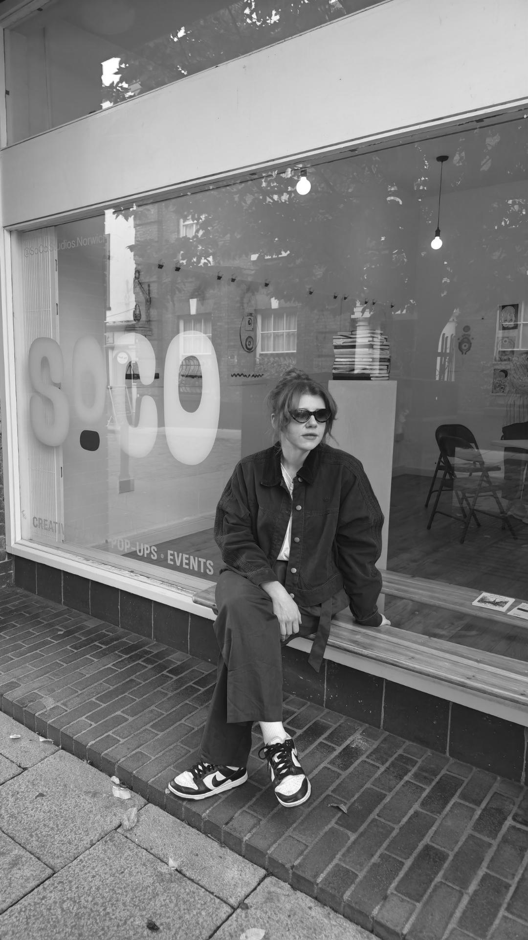 A woman sitting on a wooden bench outside a storefront window, wearing sunglasses, a dark jacket, gray pants, and sneakers.