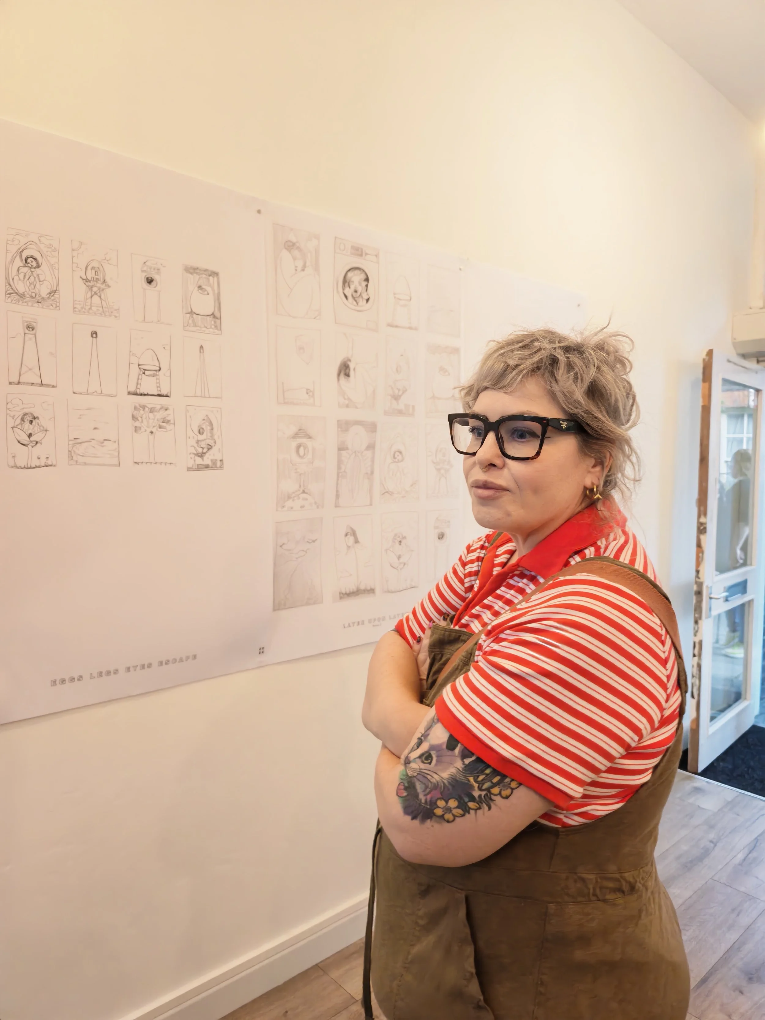 A woman with glasses and short curly gray hair wearing a red and white striped shirt and a brown apron, standing with her arms crossed, looking at a wall with sketches or drawings in an art gallery or studio.