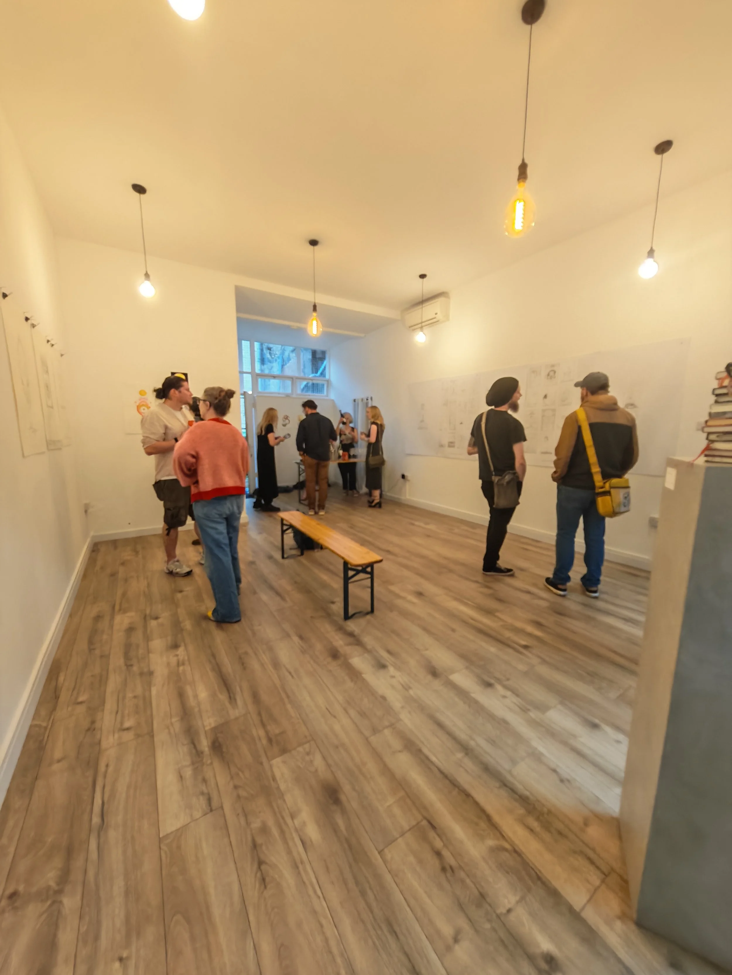 People gathered in an art gallery, some engaged in conversation, with sketches on the walls, wooden flooring, and hanging light bulbs.
