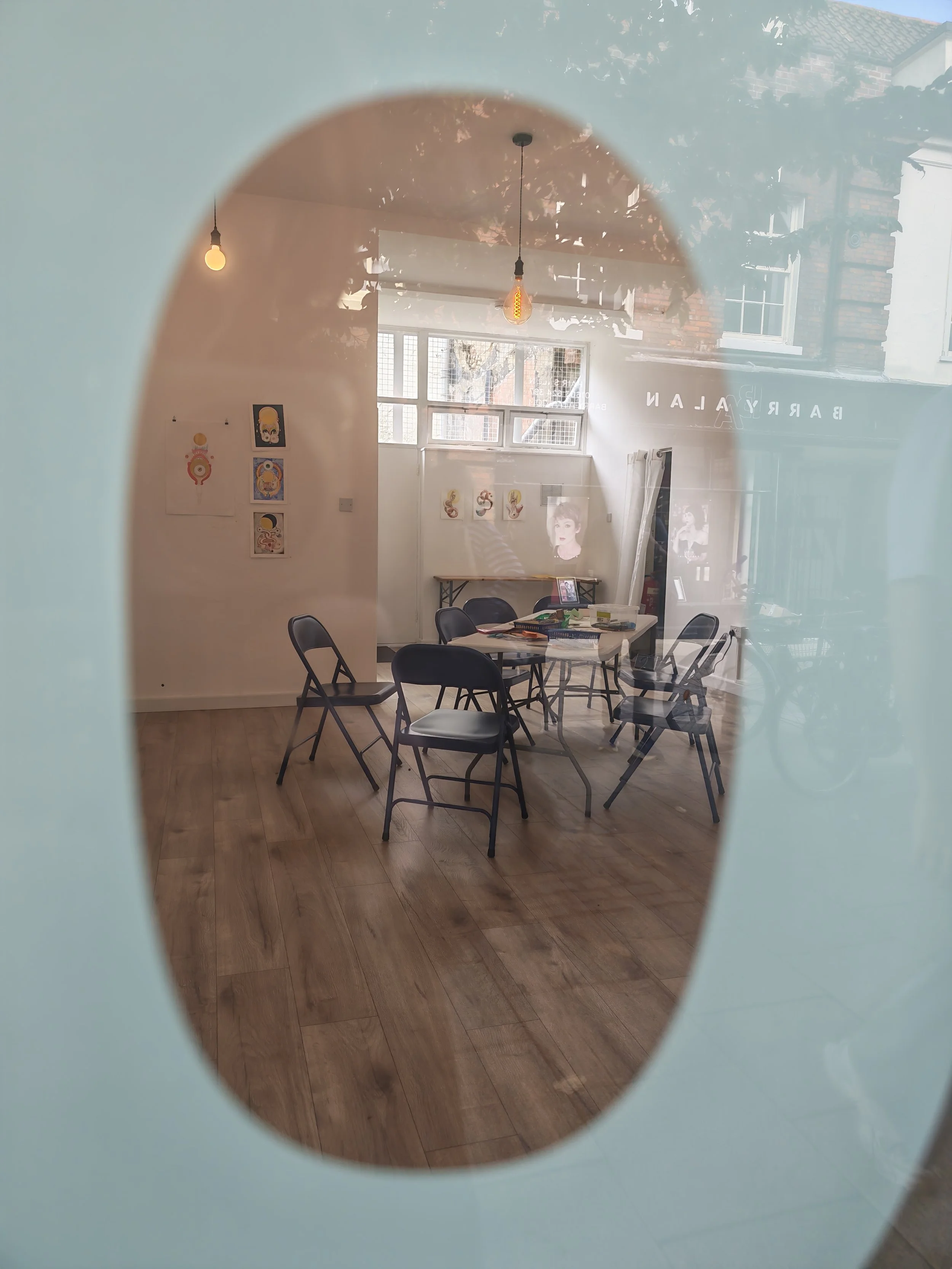 Interior of an art gallery featuring multiple framed artworks on white walls, a table with chairs, and natural light coming through a window, viewed through a circular opening in a partition.