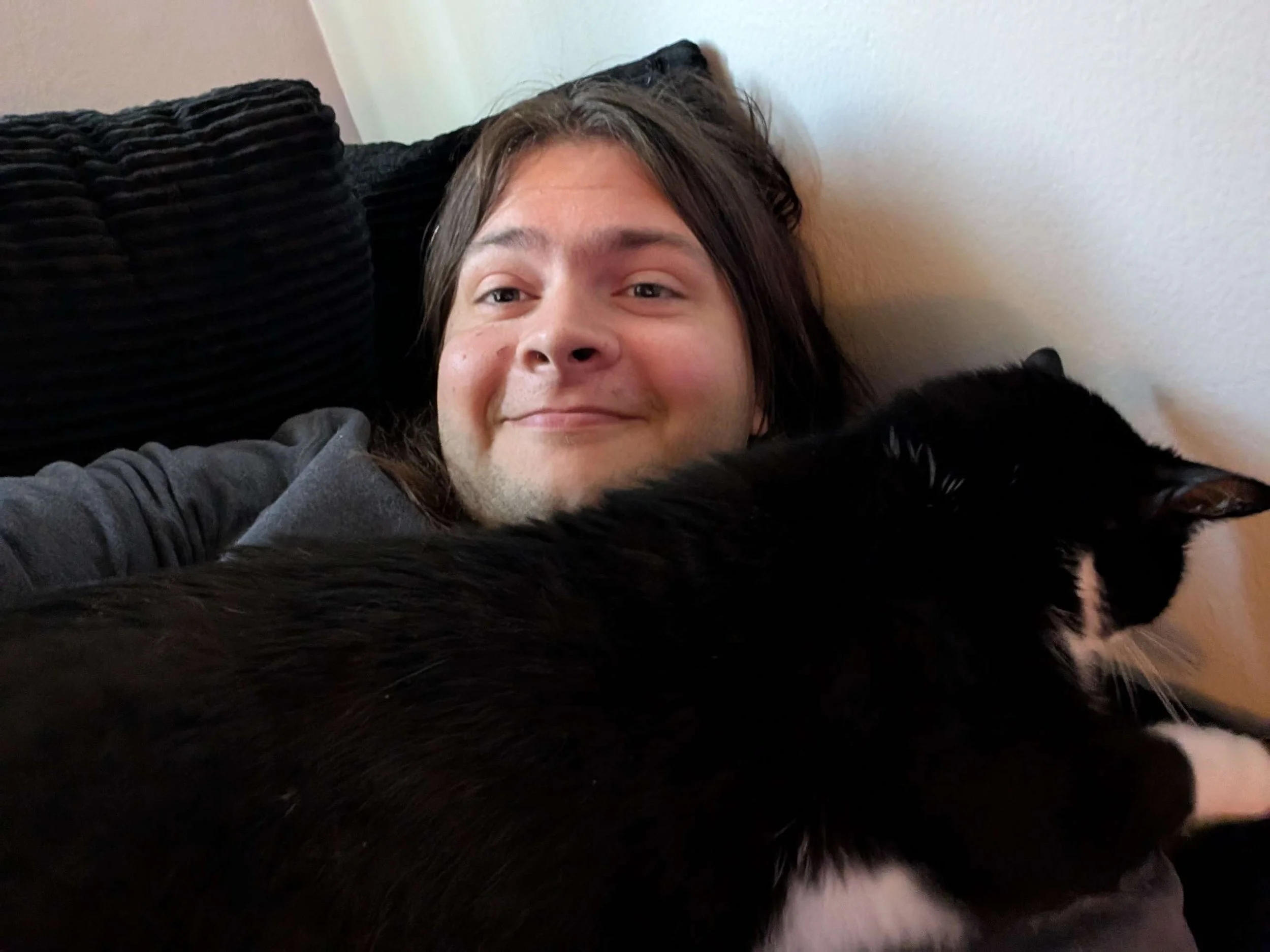 A person with long hair smiling and lying down on a black pillow, with a black and white cat in front of their face.