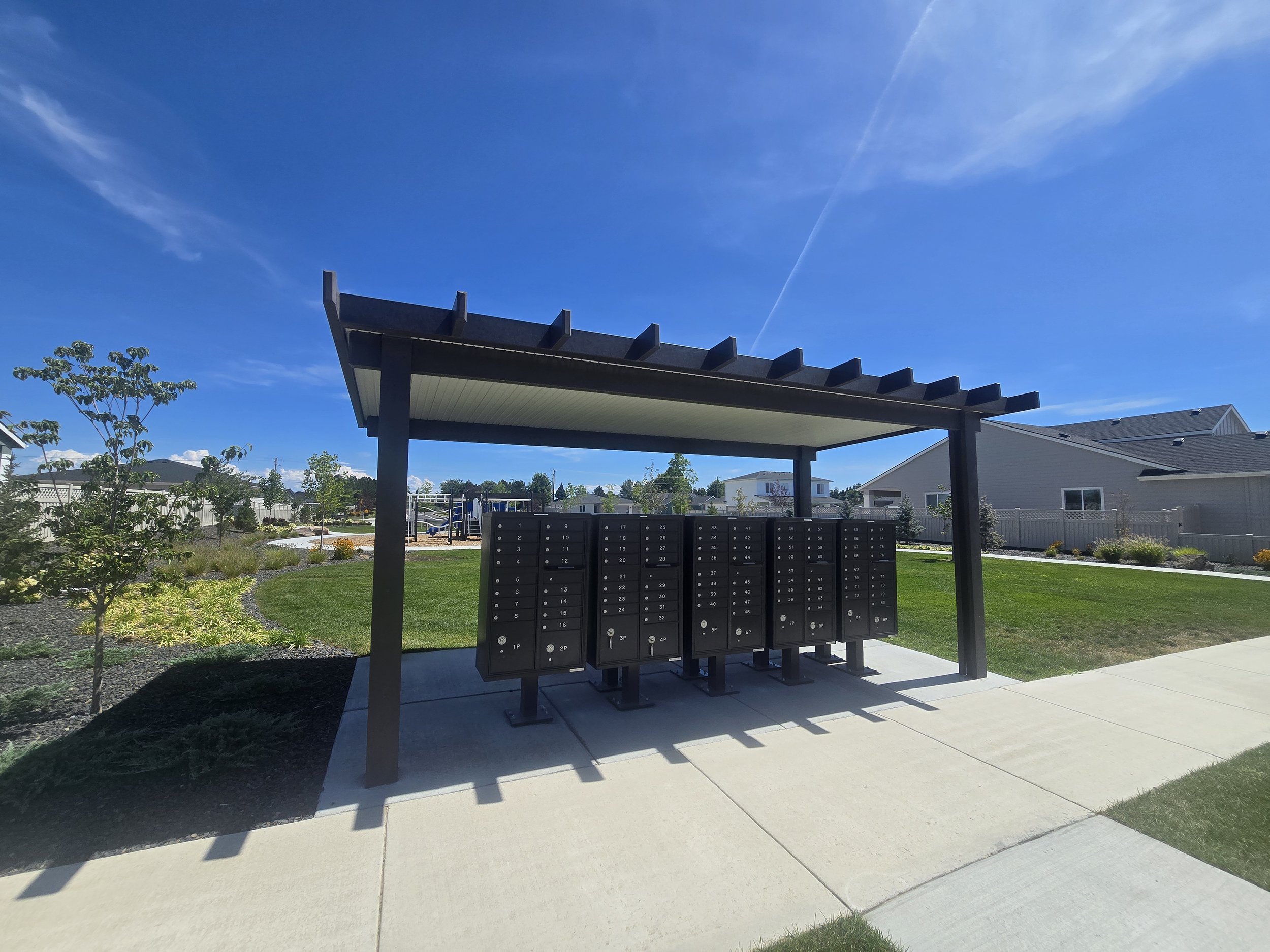 Mailboxes in a neighborhood park with a overhanging shelter, trees, houses, and a playground in the background under a clear blue sky.