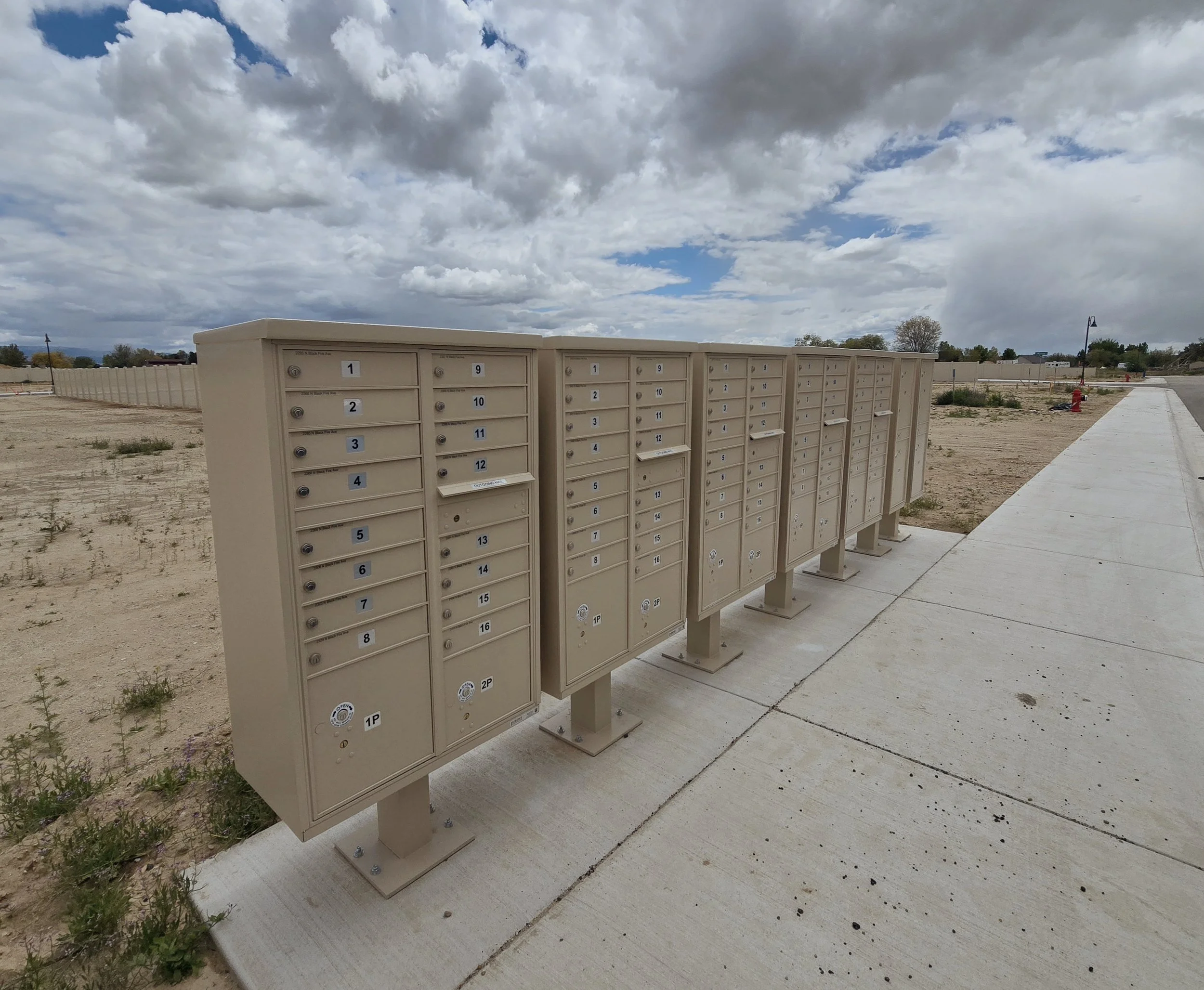 A row of beige CBU mailboxes standing outside on a concrete surface with a partly cloudy sky overhead.
