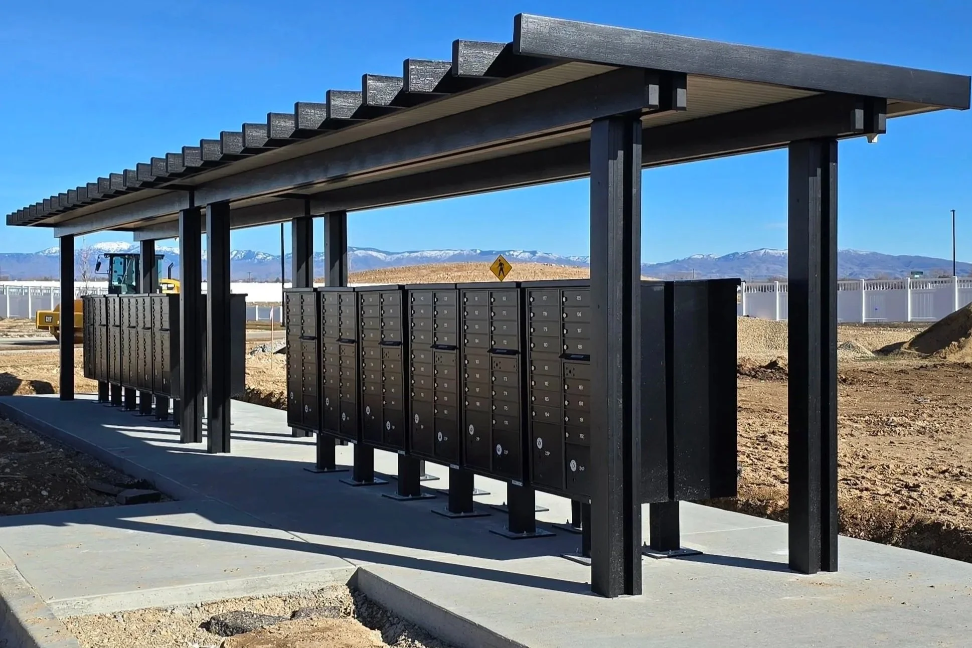 A modern outdoor mail station with multiple black mailboxes under a black roof structure, located on a concrete pad. In the background, mountains with snow and a construction machine are visible. The scene is set against a clear blue sky.