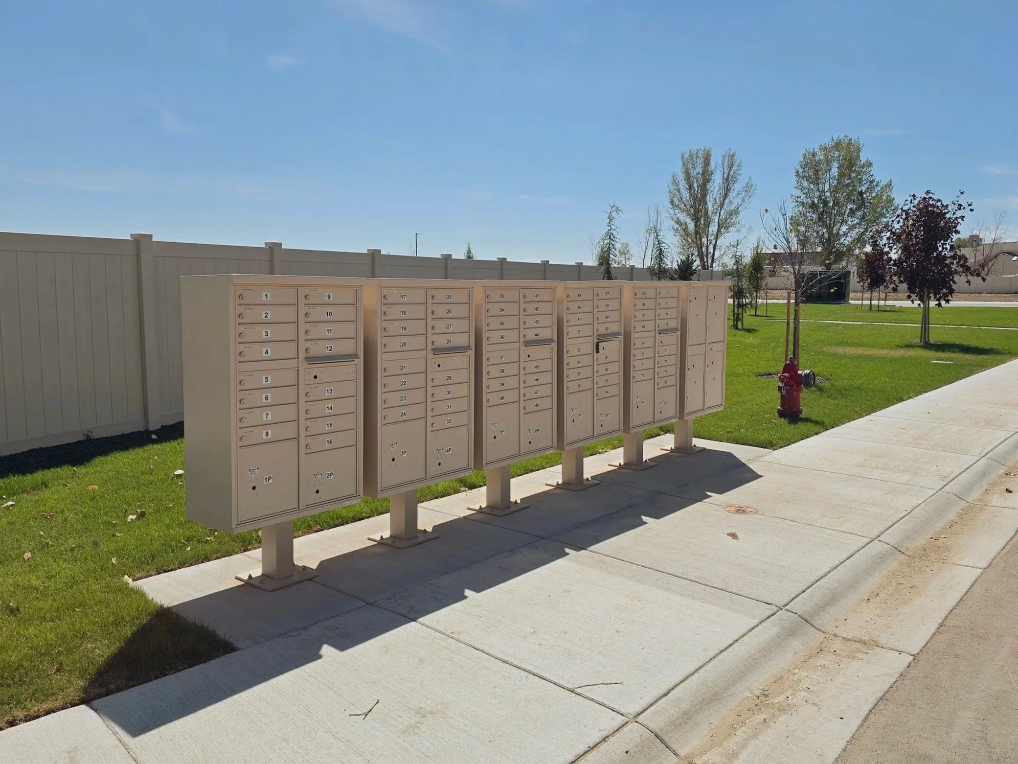 A row of beige mailboxes outdoors on a sunny day, with a white fence, green grass, and trees in the background.