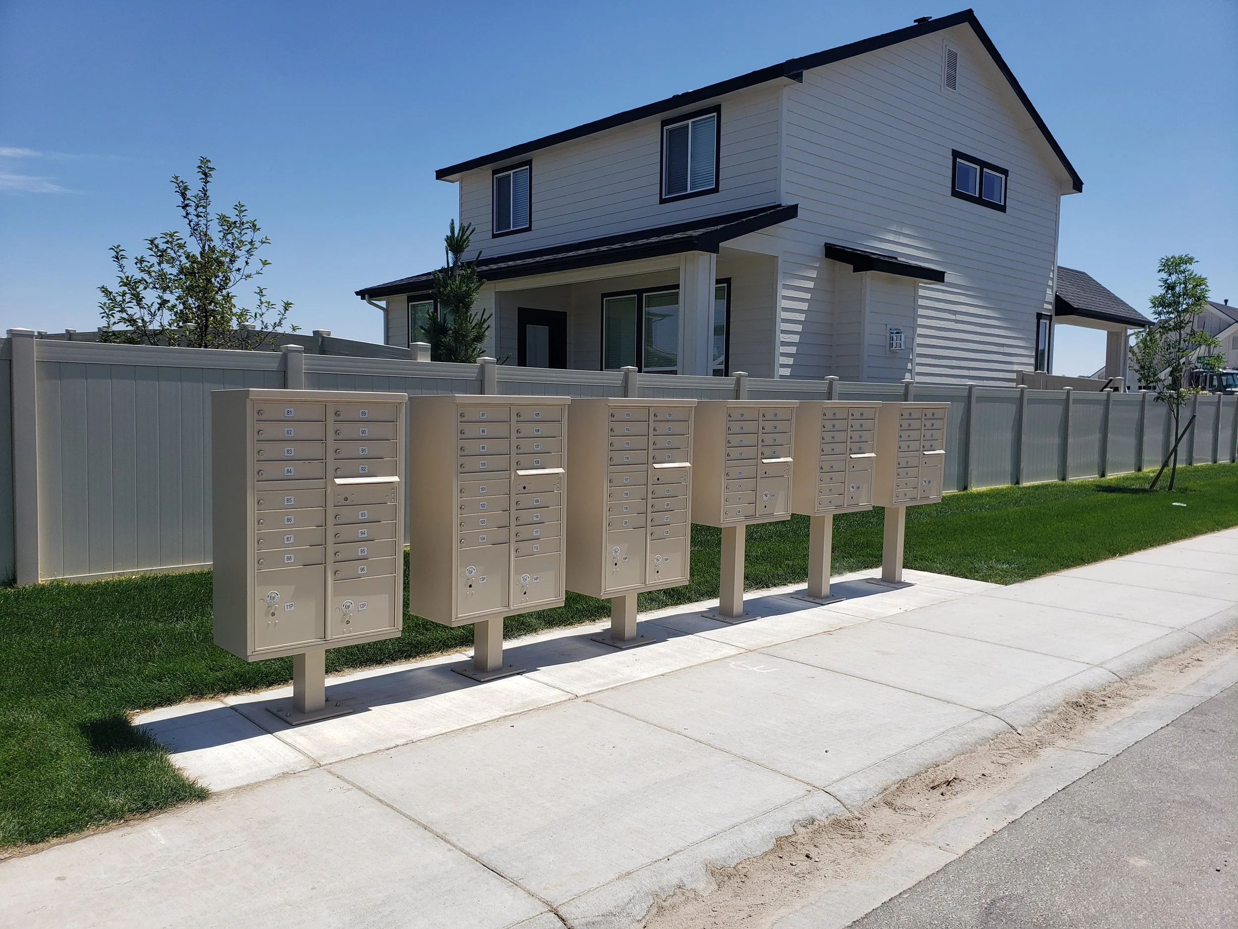 A row of beige CBU mailboxes stands on a sidewalk in front of a green lawn. In the background, a white two-story house with black accents and windows is visible, with a blue sky overhead.