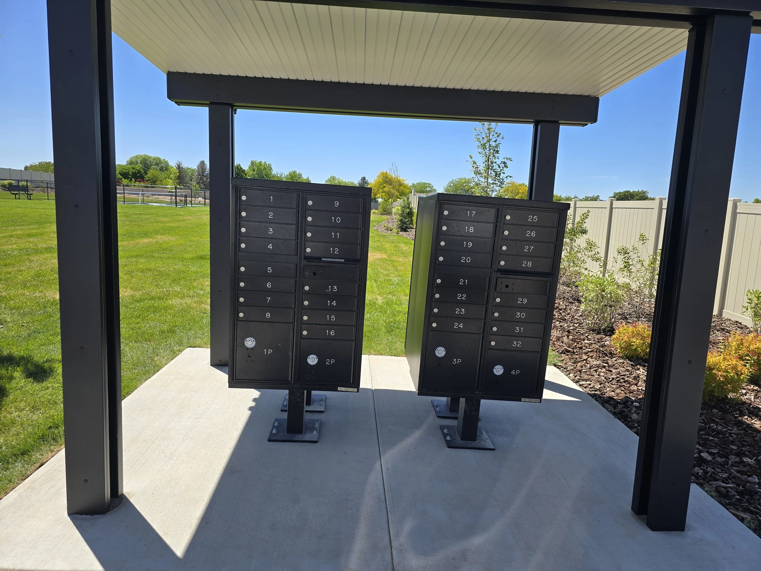 A postal mailbox station with multiple black metal mailboxes numbered from 1 to 32, installed under a sheltered structure with a beige roof and supported by black metal posts, set on a concrete slab in a grassy park area with trees and a fence in the background during sunny weather.