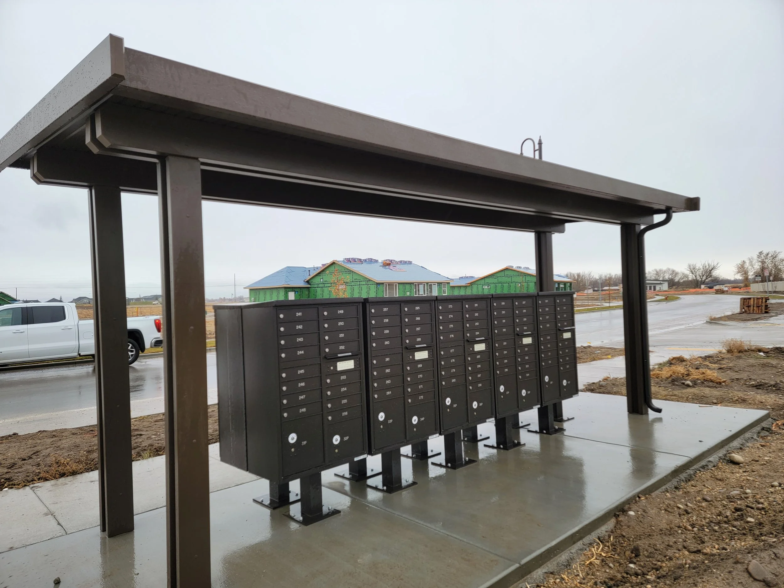 View of a mailbox area under a metal roof structure with multiple mailboxes, with a house under construction and a car in the background during cloudy weather.