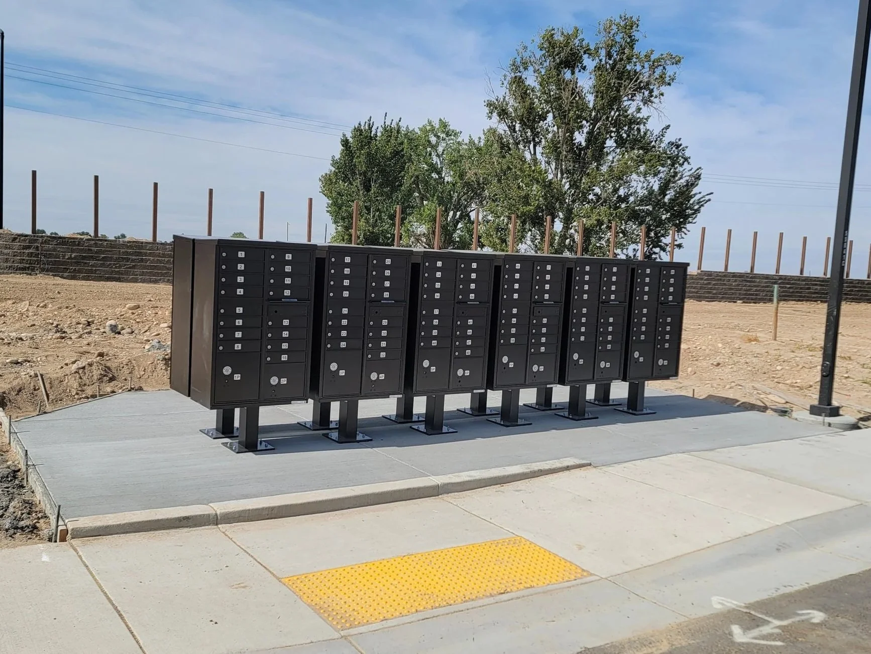 A row of black cluster box units (CBU)mailboxes with numbers near a sidewalk in an outdoor area, with trees and a blue sky in the background.