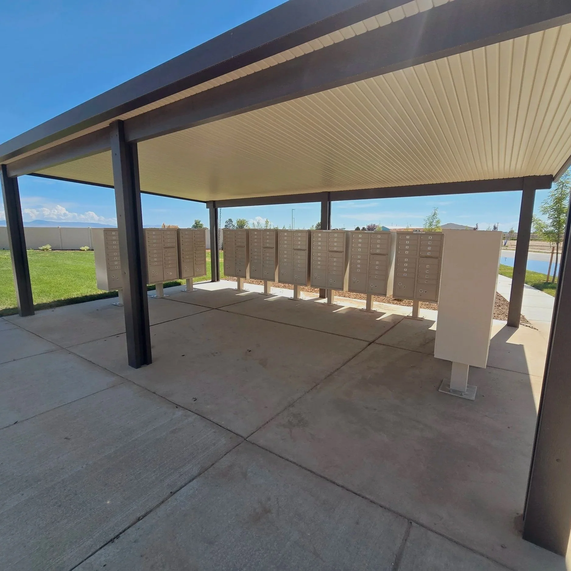 This image shows an outdoor mail collection station with multiple beige mailboxes arranged in rows and a covered roof structure overhead. There is a sidewalk and some greenery around the area.