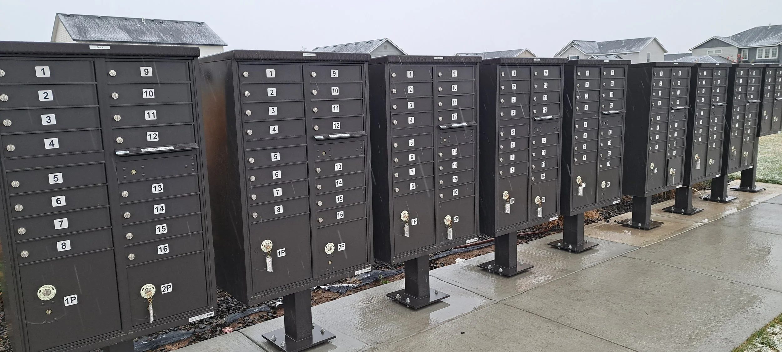 Row of black CBU mailboxes with combination locks and numbered labels, situated on a sidewalk in a residential area with houses in the background and light snow on the rooftops.