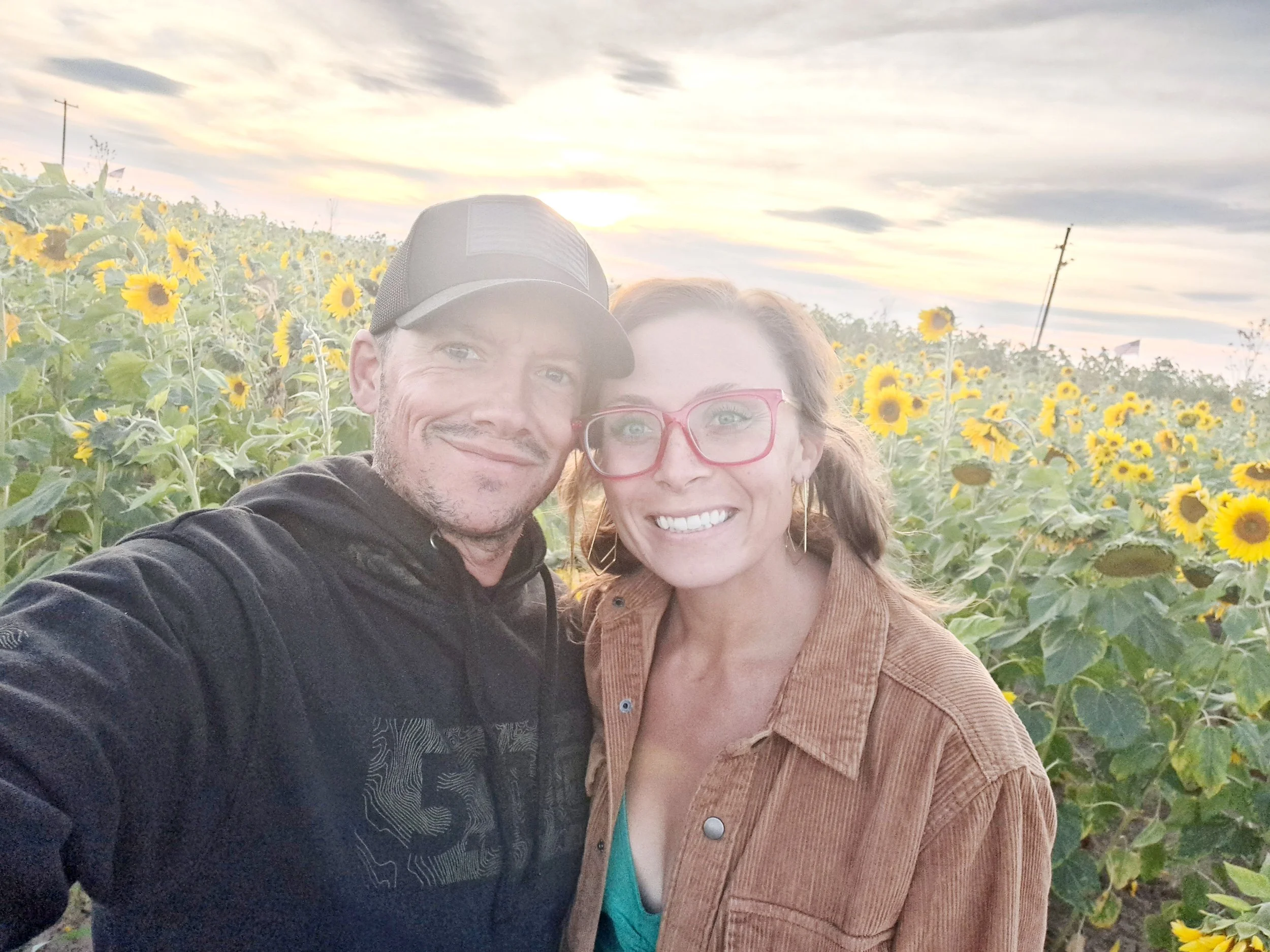 A smiling couple takes a selfie in a sunflower field at sunset, with cloudy sky and power lines in the background.