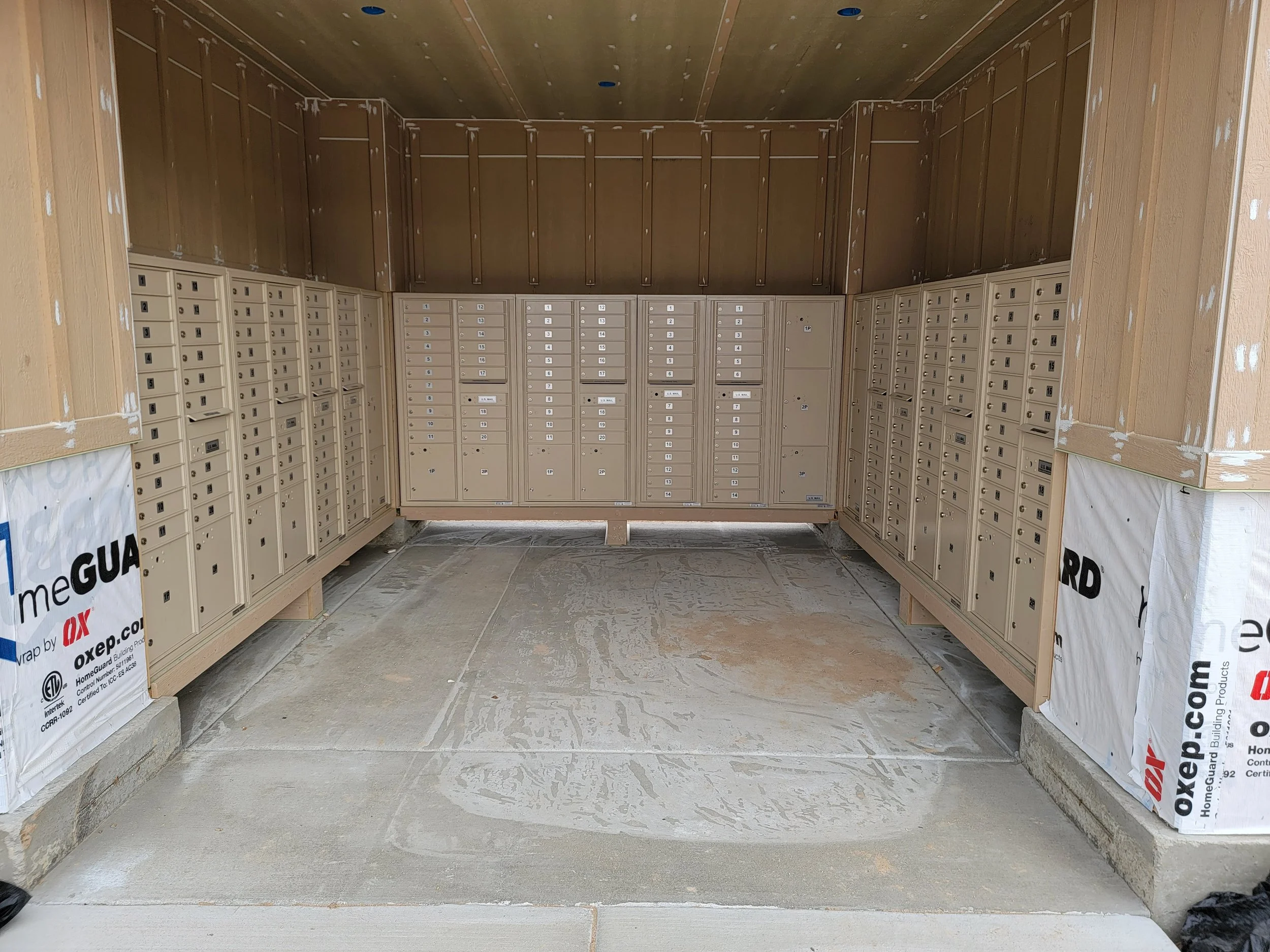 Large covered area with beige mailboxes on both sides, under construction with unfinished drywall and insulation visible, concrete floor, and patchwork on walls and ceiling.