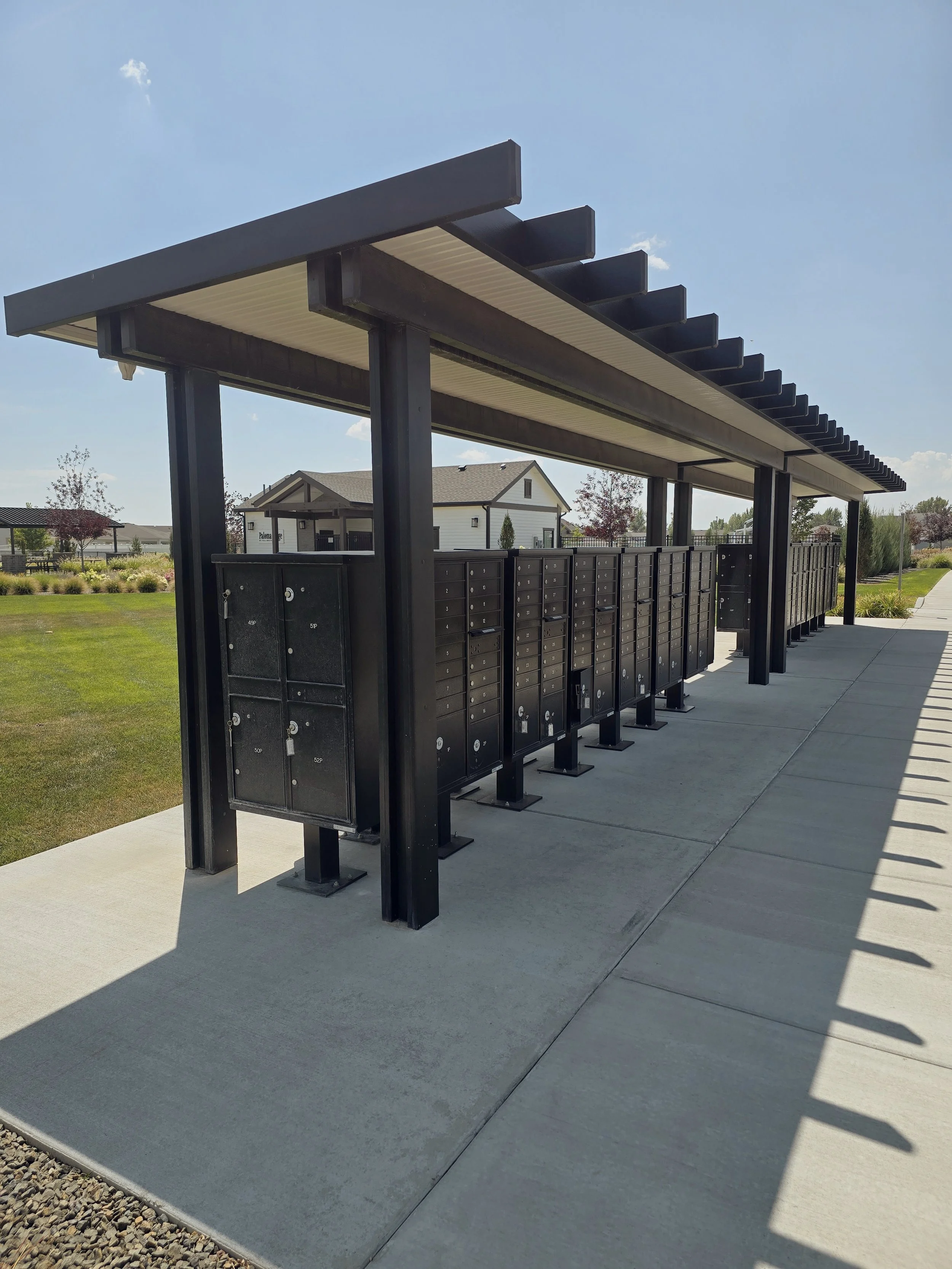 Row of black cluster box units (CBU)mailboxes under a covered shelter with a white and gray residential house in the background, set on a concrete sidewalk with grassy areas and small trees nearby.