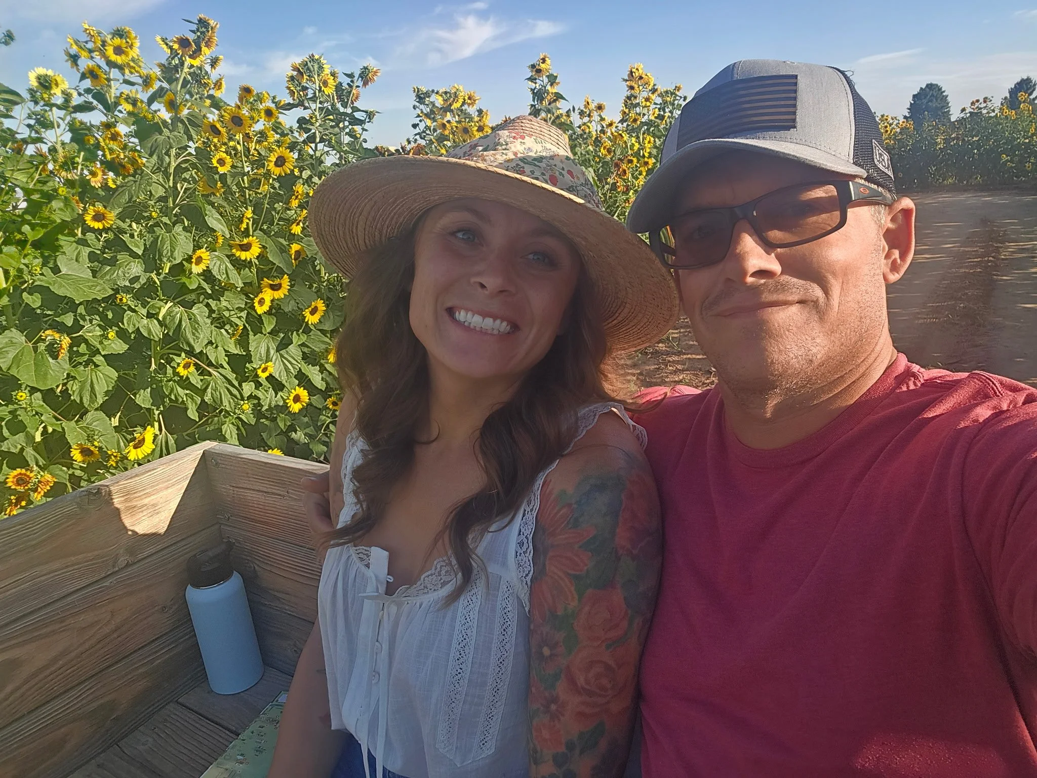 A smiling woman and man taking a selfie together in a sunflower field on a sunny day. The woman has long dark hair and is wearing a large straw hat and a white top. The man is wearing glasses and a gray cap. There is a wooden bench and a water bottle visible in the background.