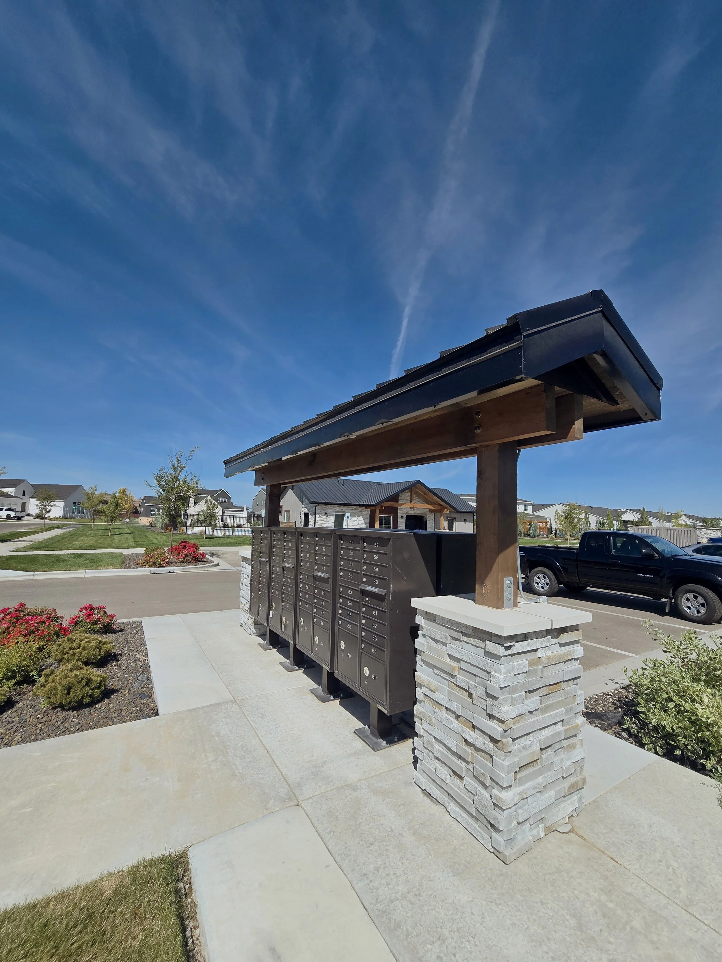 A mail kiosk with multiple compartments under a small roof structure, situated in a landscaped residential area with houses, trees, and a parked black truck in the background during a sunny day.