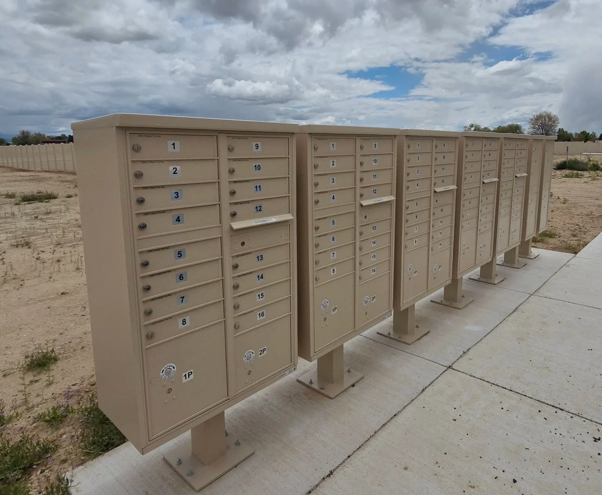 Multiple beige mailboxes in an outdoor setting with a cloudy sky overhead.