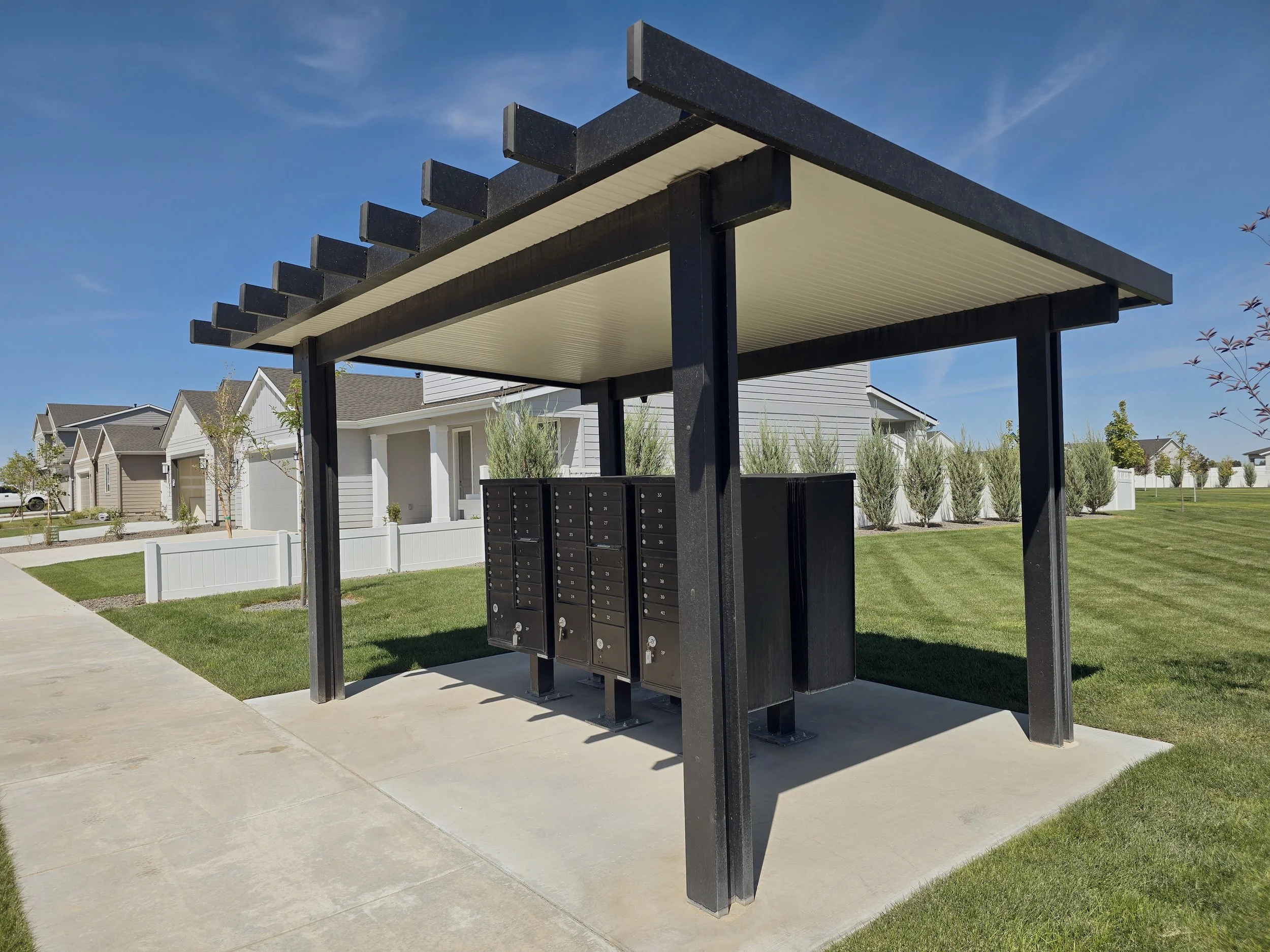 A modern outdoor mailbox station with multiple mailboxes under a black pergola in a suburban neighborhood.