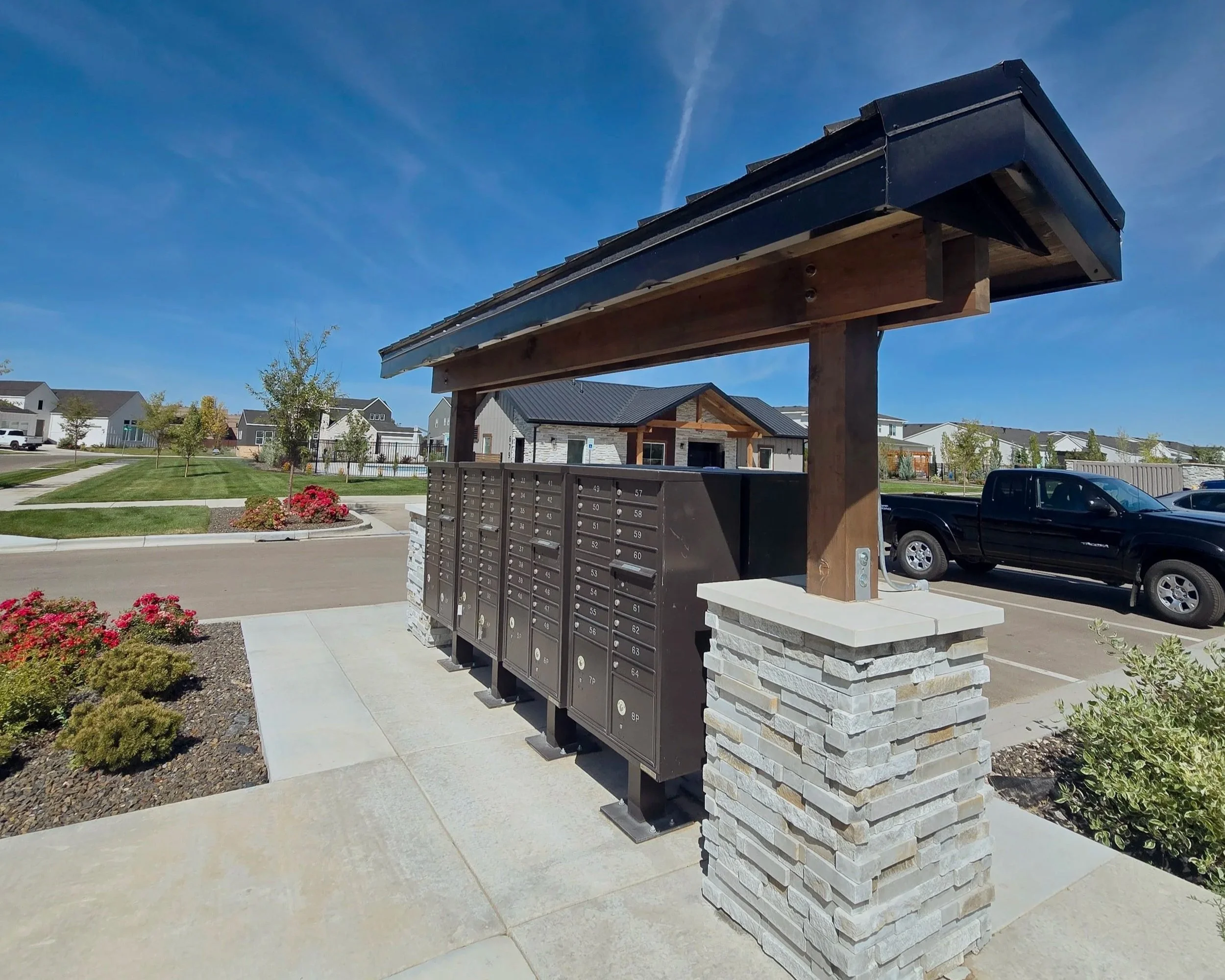 Apartment complex with a row of mailboxes in front, a small landscaped area with flowers and shrubs, and a blue sky with some clouds.