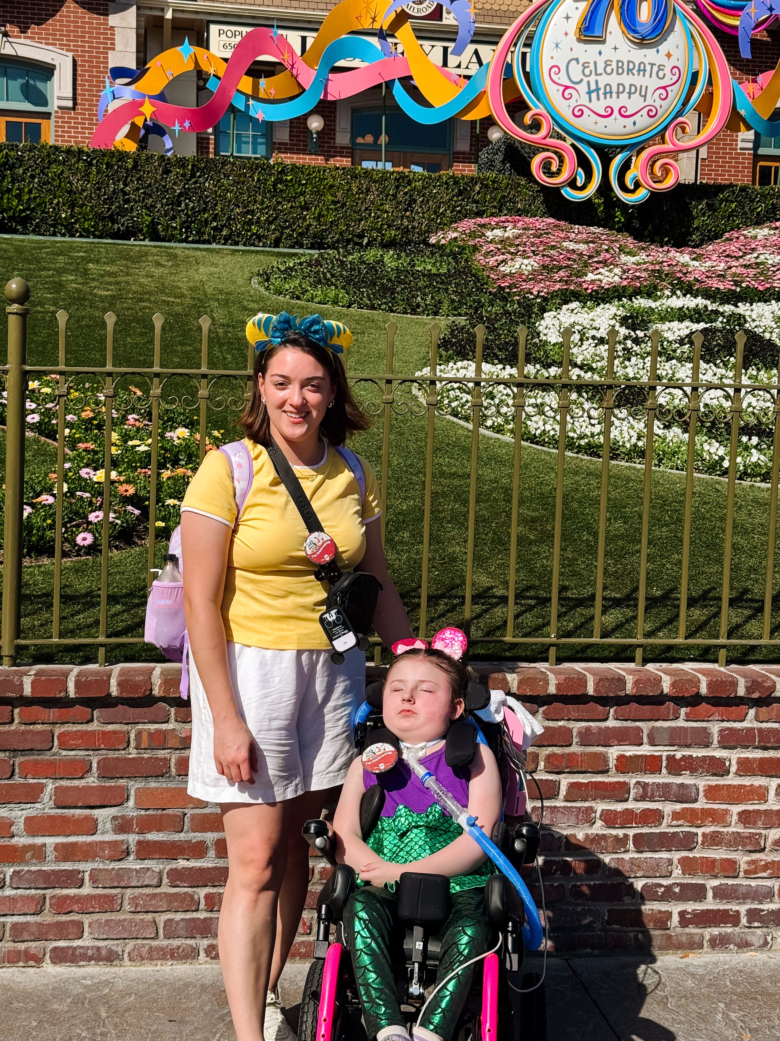 A woman with a yellow shirt standing next to Awty who is in a wheelchair, in front of a Disneyland sign