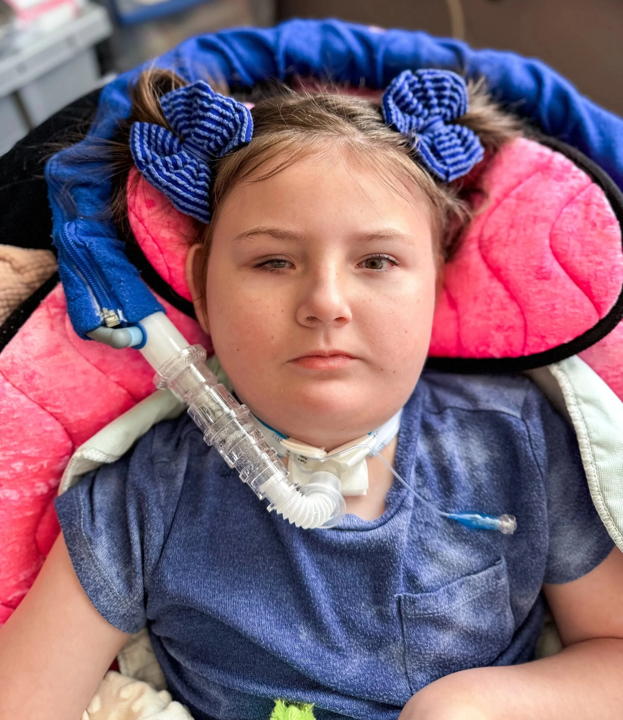 A photo of Awty from the torso up, wearing a blue shirt, blue hair bows, and laying in a pink beanbag with her ventilator hooked up.