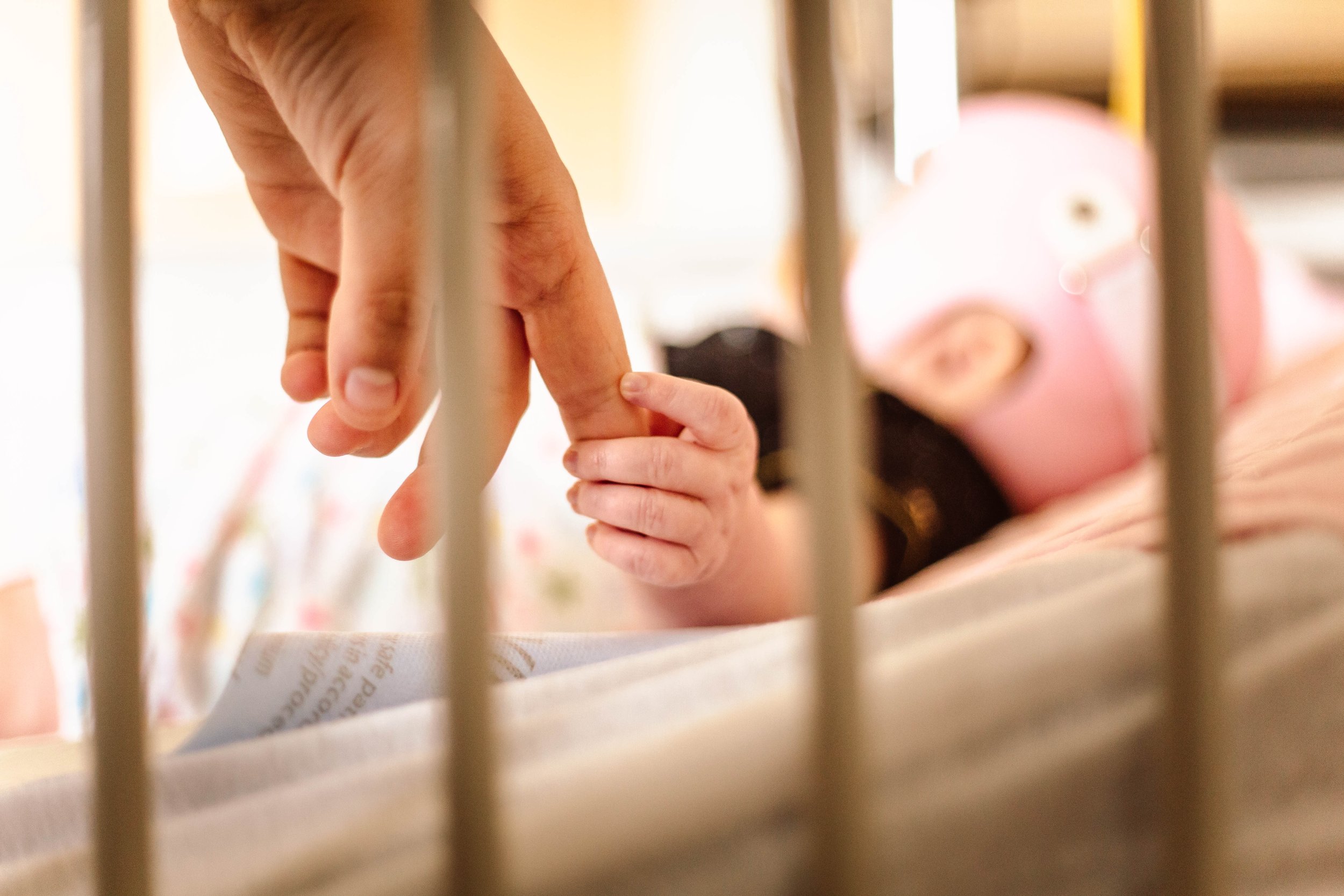 An adult hand in focus behind the bars of a hospital crib, being held by a baby hand, with the baby blurry in the background with a medical helmet on
