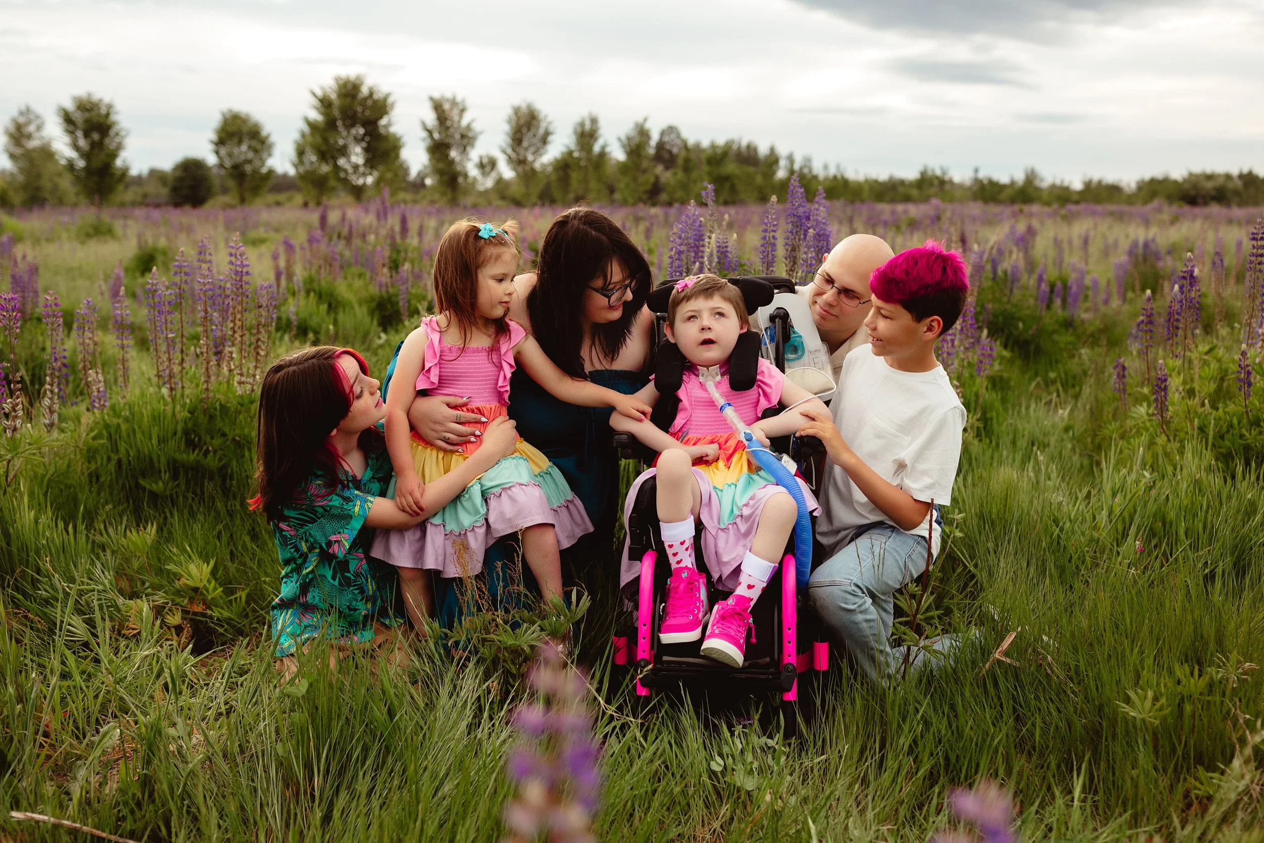 Awty in a pink wheelchair and bright dress in the middle of a field of purple lupine flowers surrounded by her parents and siblings kneeling around her.
