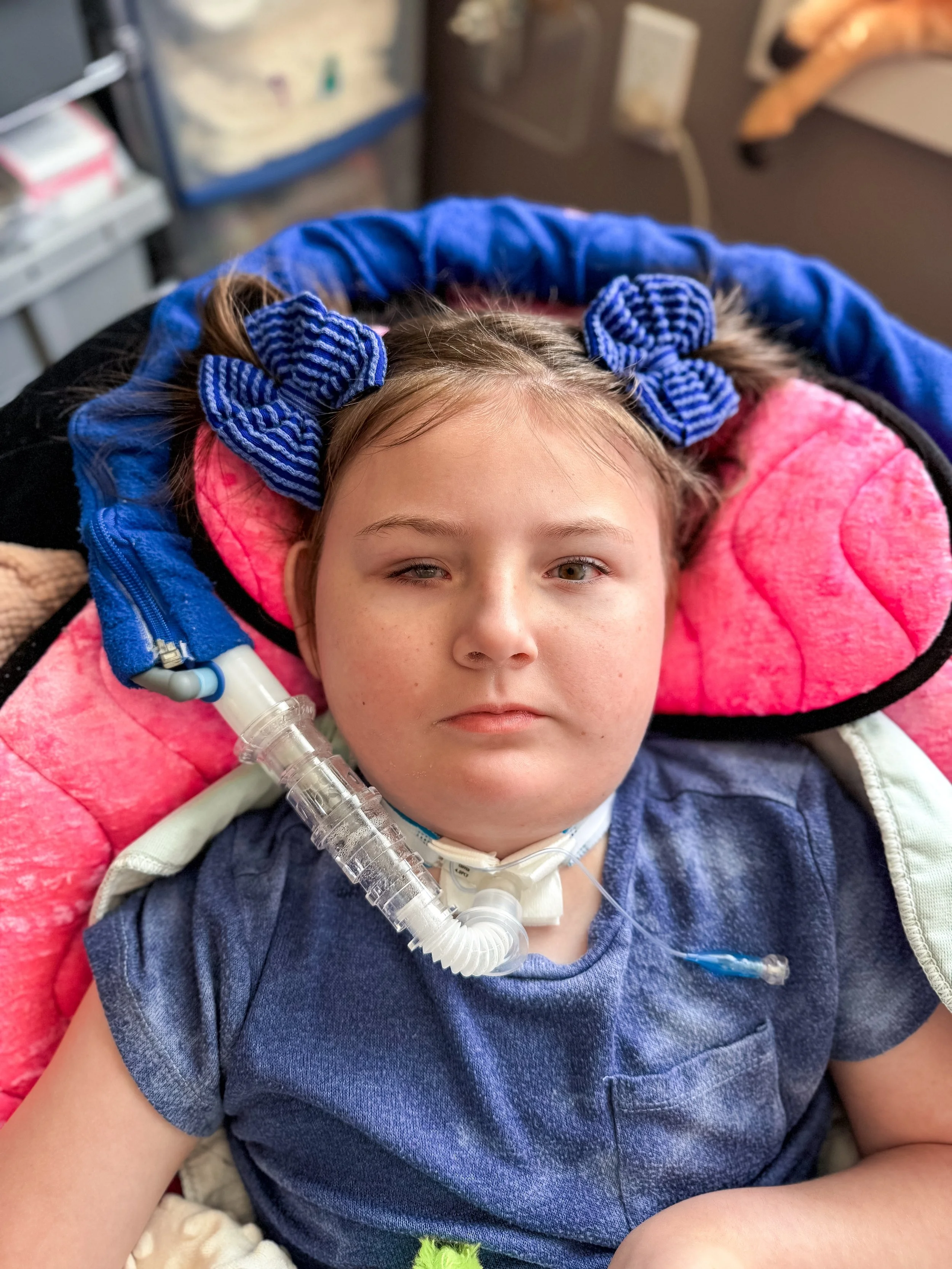 Awty from the torso up, wearing a blue shirt with blue hair bows, laying in a pink beanbag with her ventilator hooked up