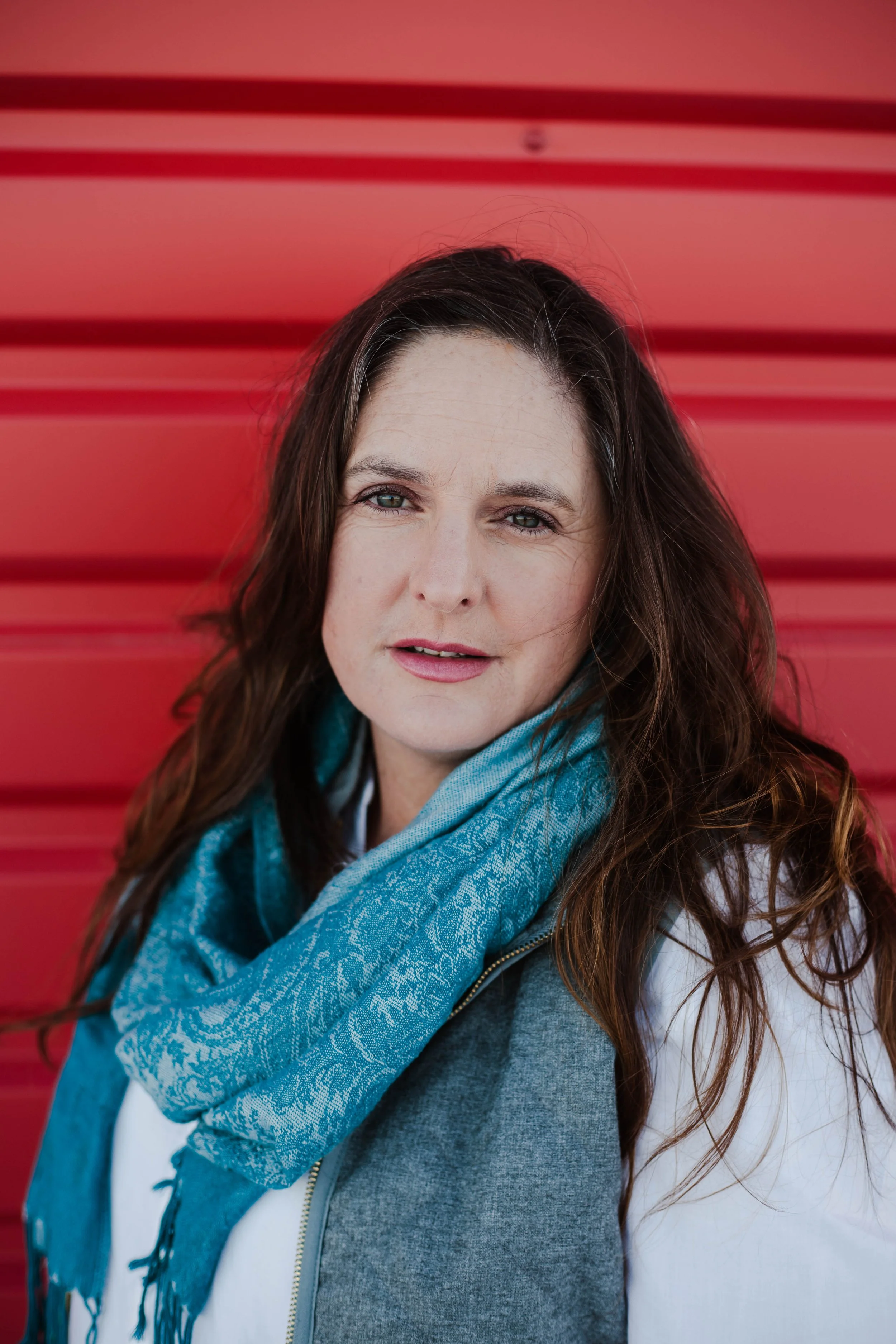 A caucasian woman from the shoulders up with dark hair and a big blue scarf in front of a red background