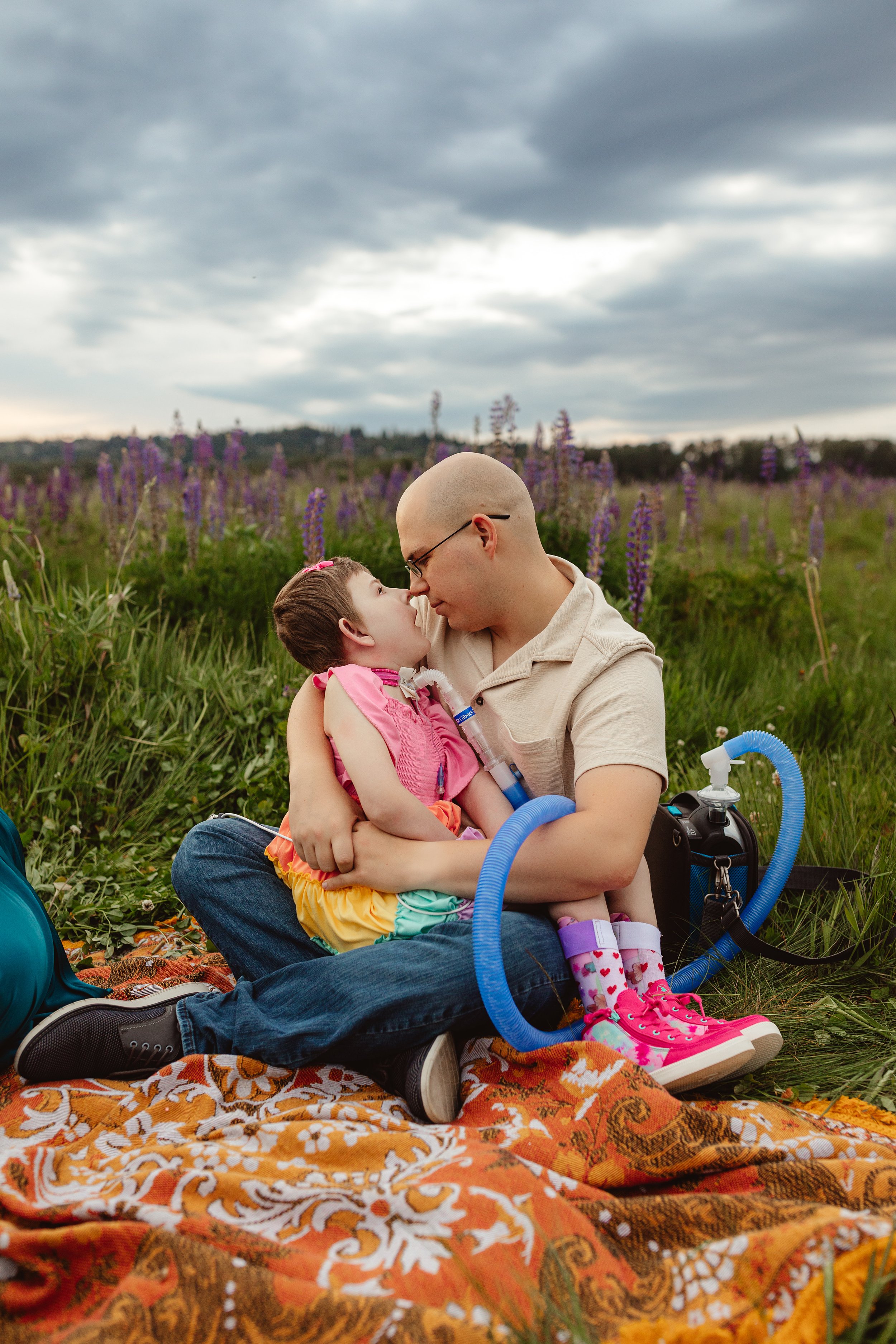 A man sitting in a field of lupine flowers holding Awty with a ventilator on the ground beside them