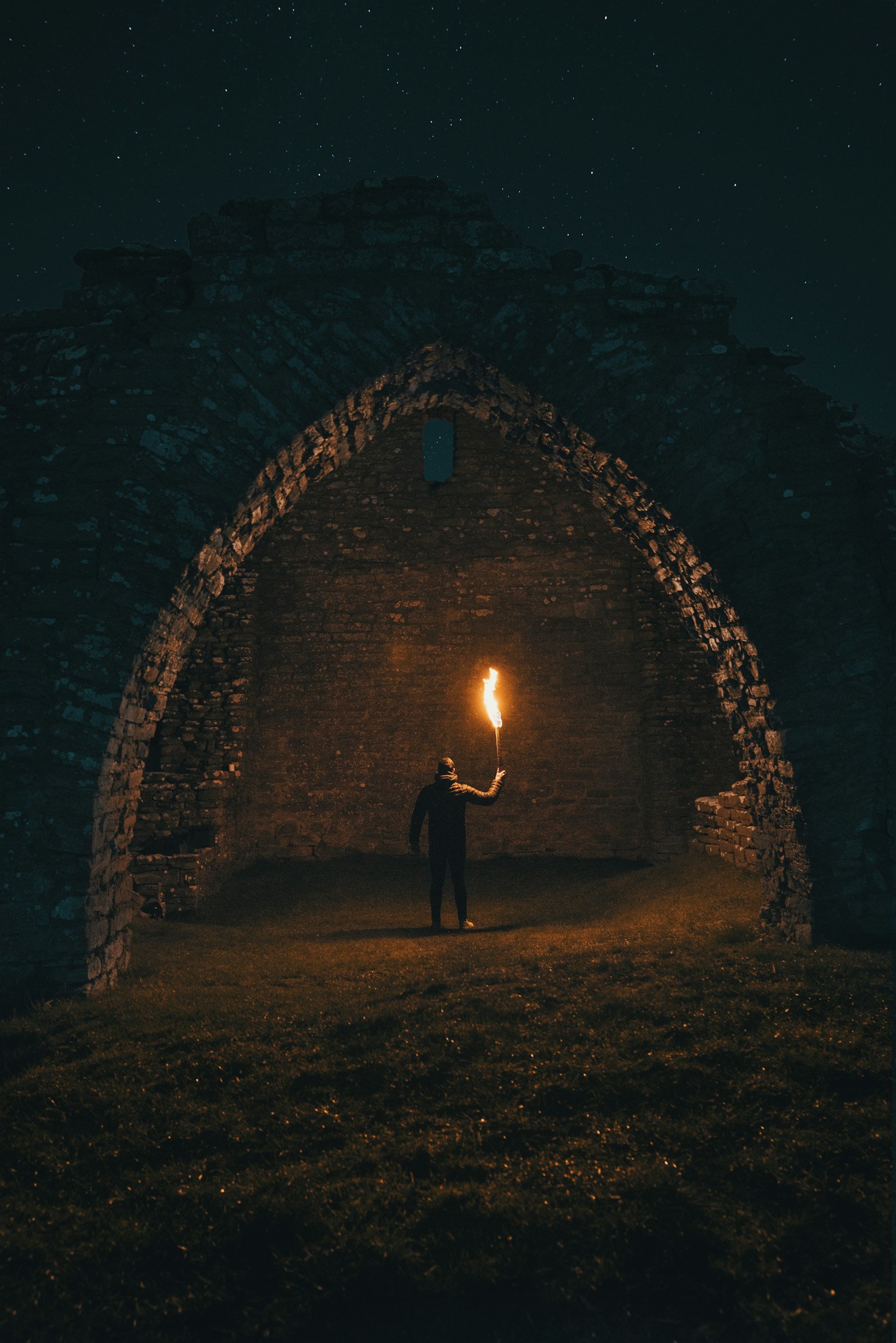 A person holding a flaming torch standing inside an ancient stone archway at night beneath a starry sky.
