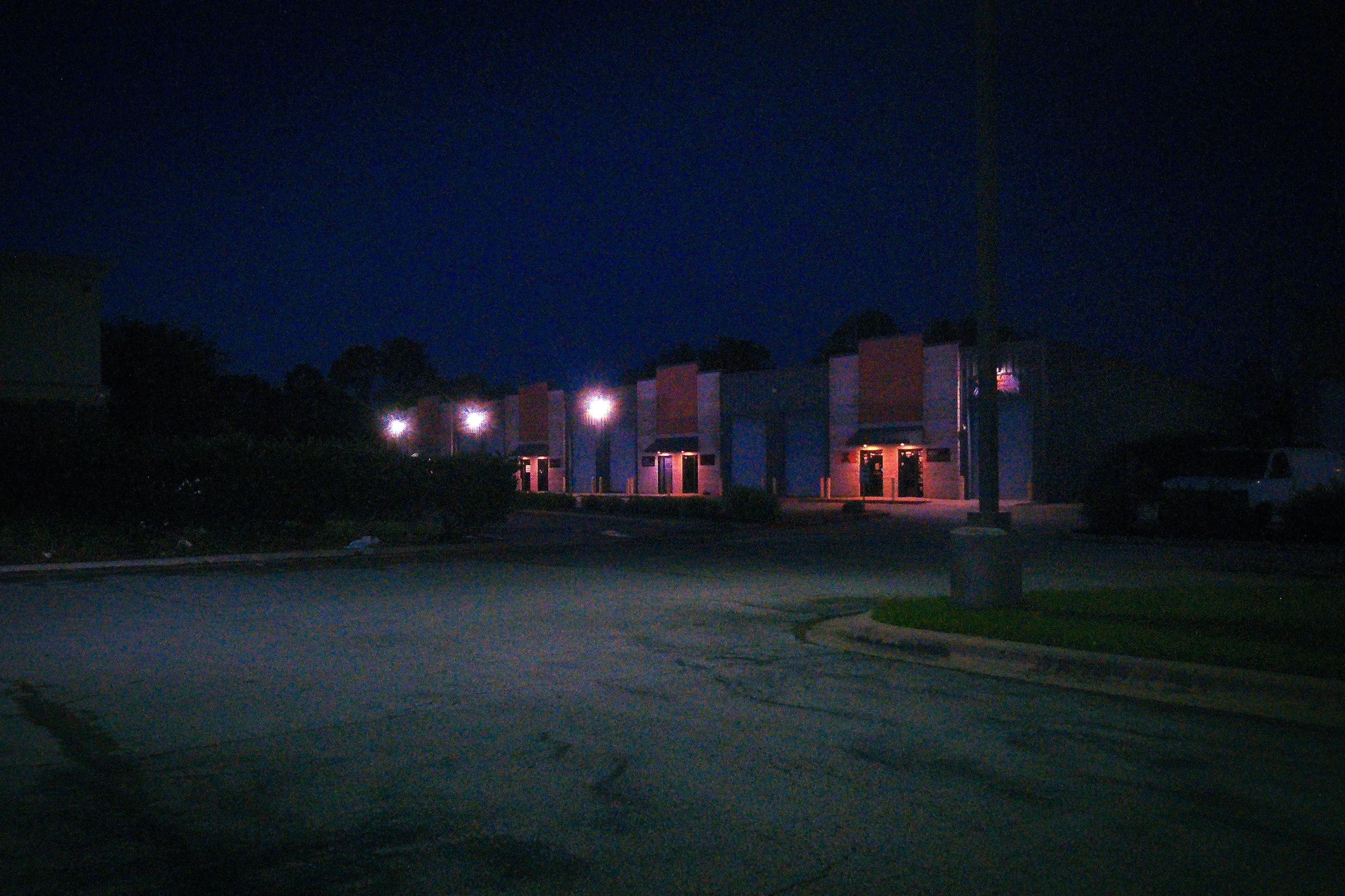Night scene of a parking lot with a row of buildings illuminated by exterior lights, dark sky, and a few parked vehicles.