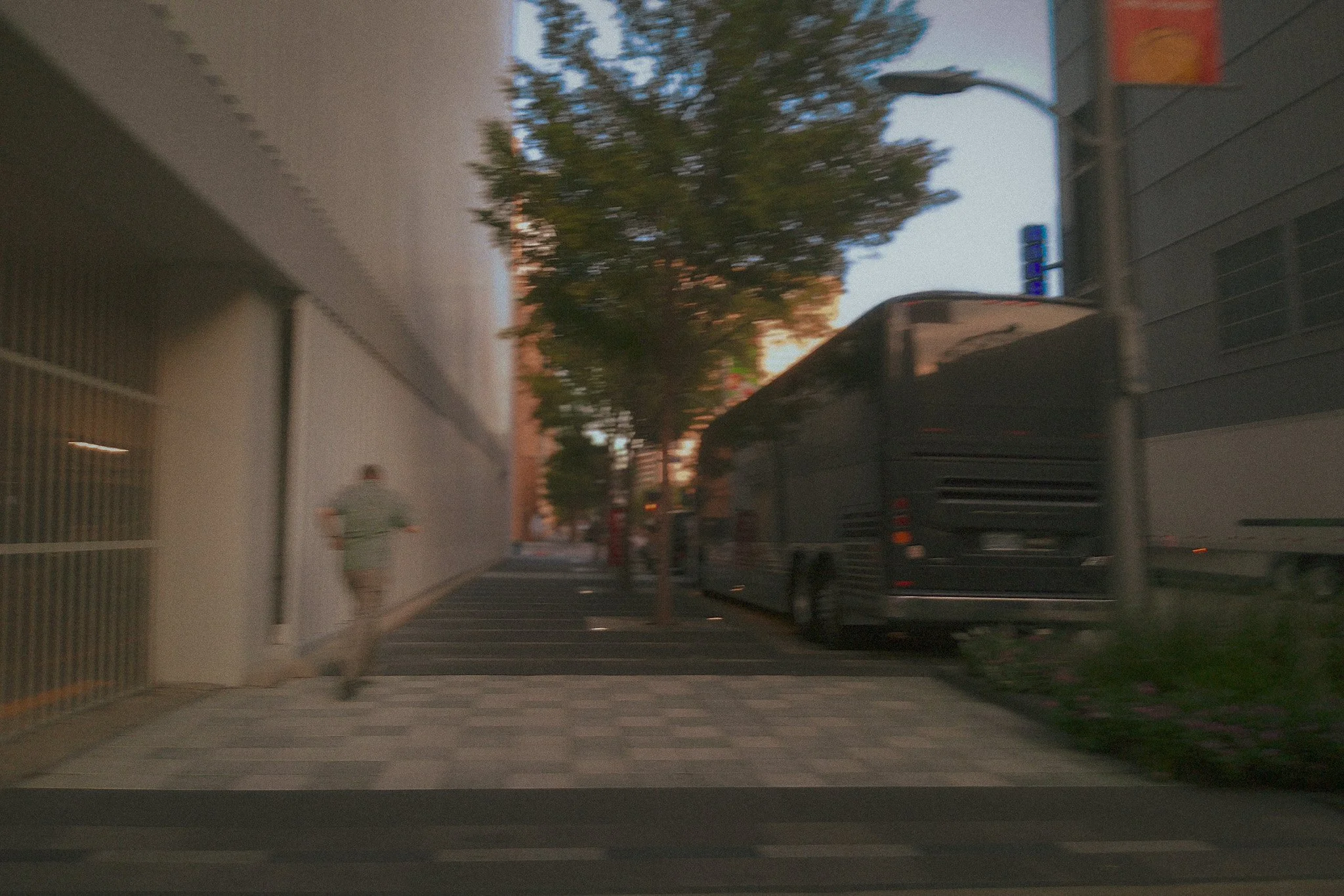 A sidewalk with a person running away, a large tree, a trash bin, a building wall on the left, and a vehicle parked on the street, all seen during dusk  with a partly cloudy sky.