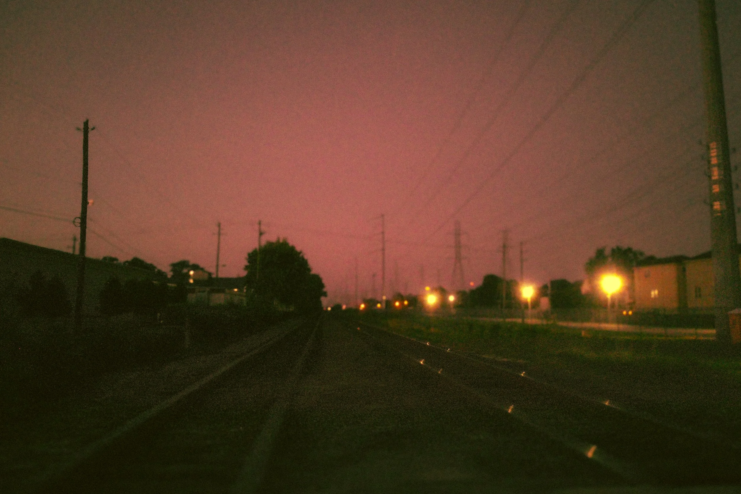 A street scene at night with a pinkish sky, utility poles and lines, houses, trees, and streetlights illuminating the street.