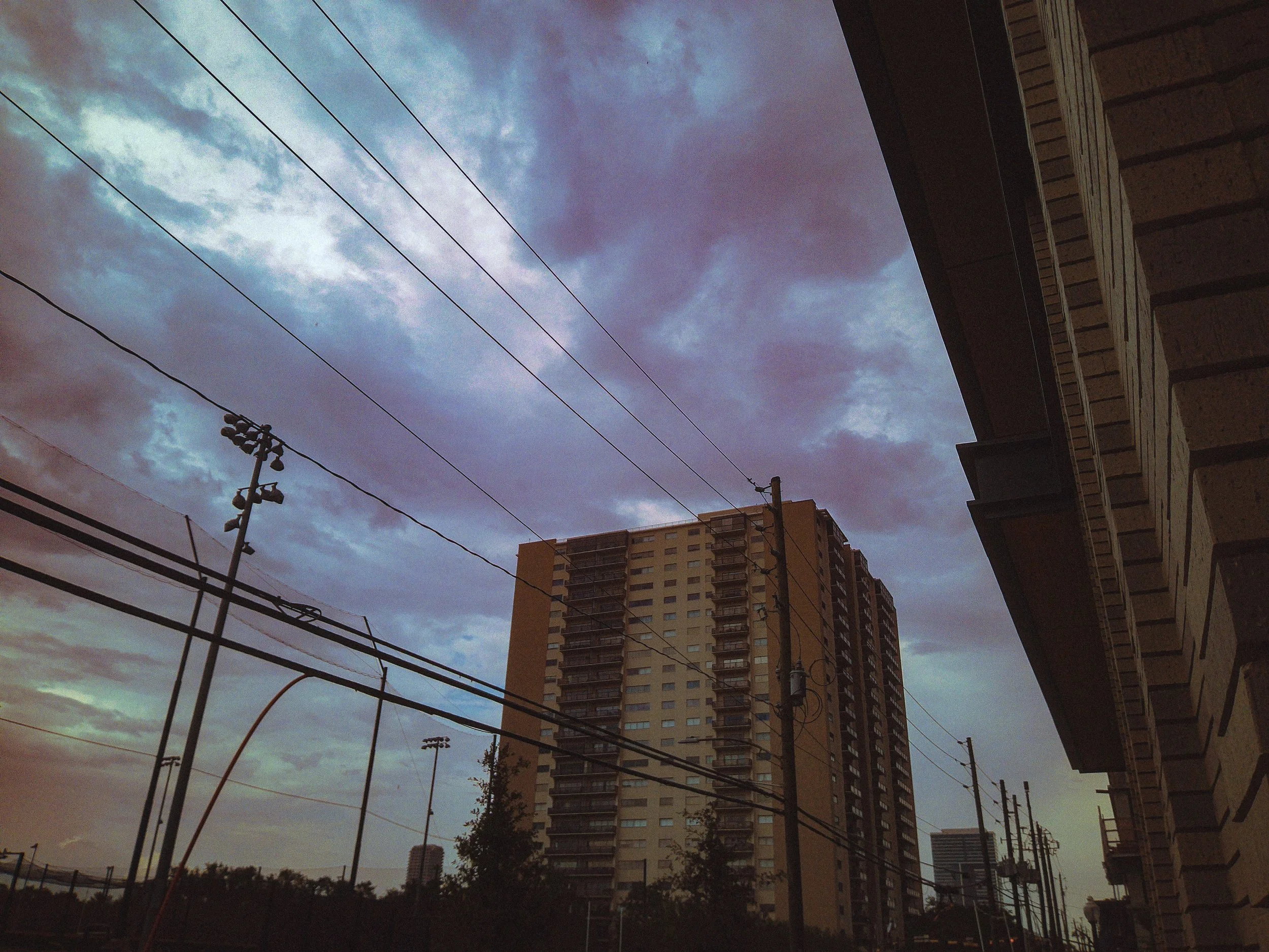 View of a cloudy sky over tall buildings and utility poles with power lines, taken from an urban area.