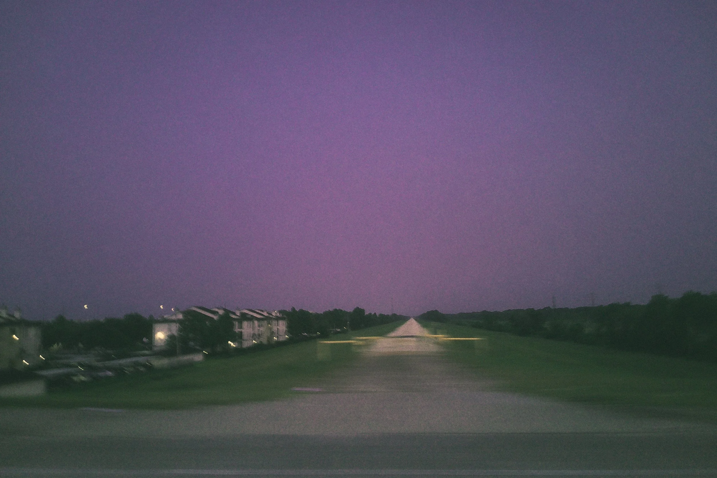 A suburban neighborhood street at dusk with a darkening sky showing shades of purple, pink, and blue, with houses on the left and a winding road with green grass in the foreground.