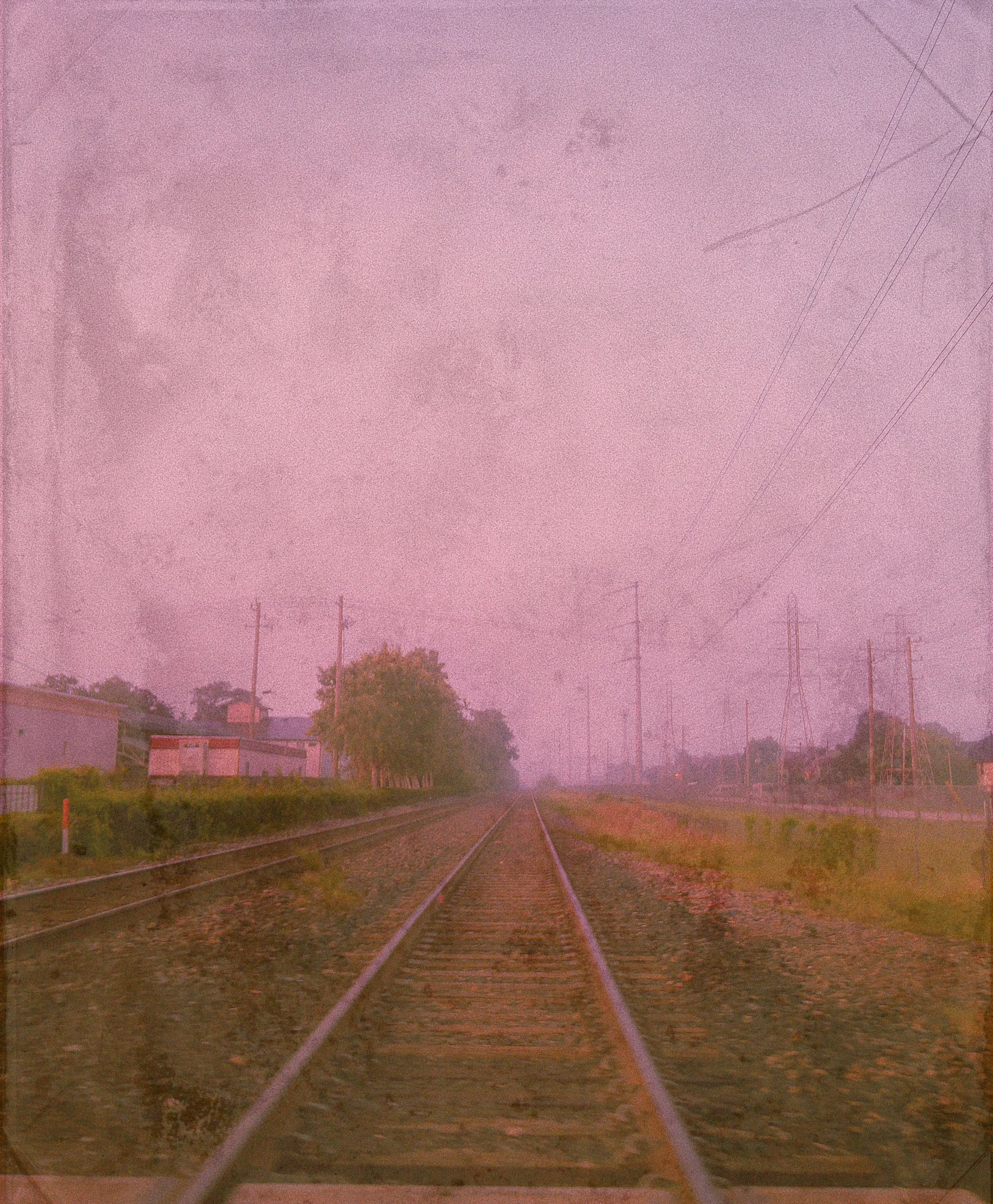 A set of railroad tracks running through rural land under a pink or purple sky with power lines and trees in the background.