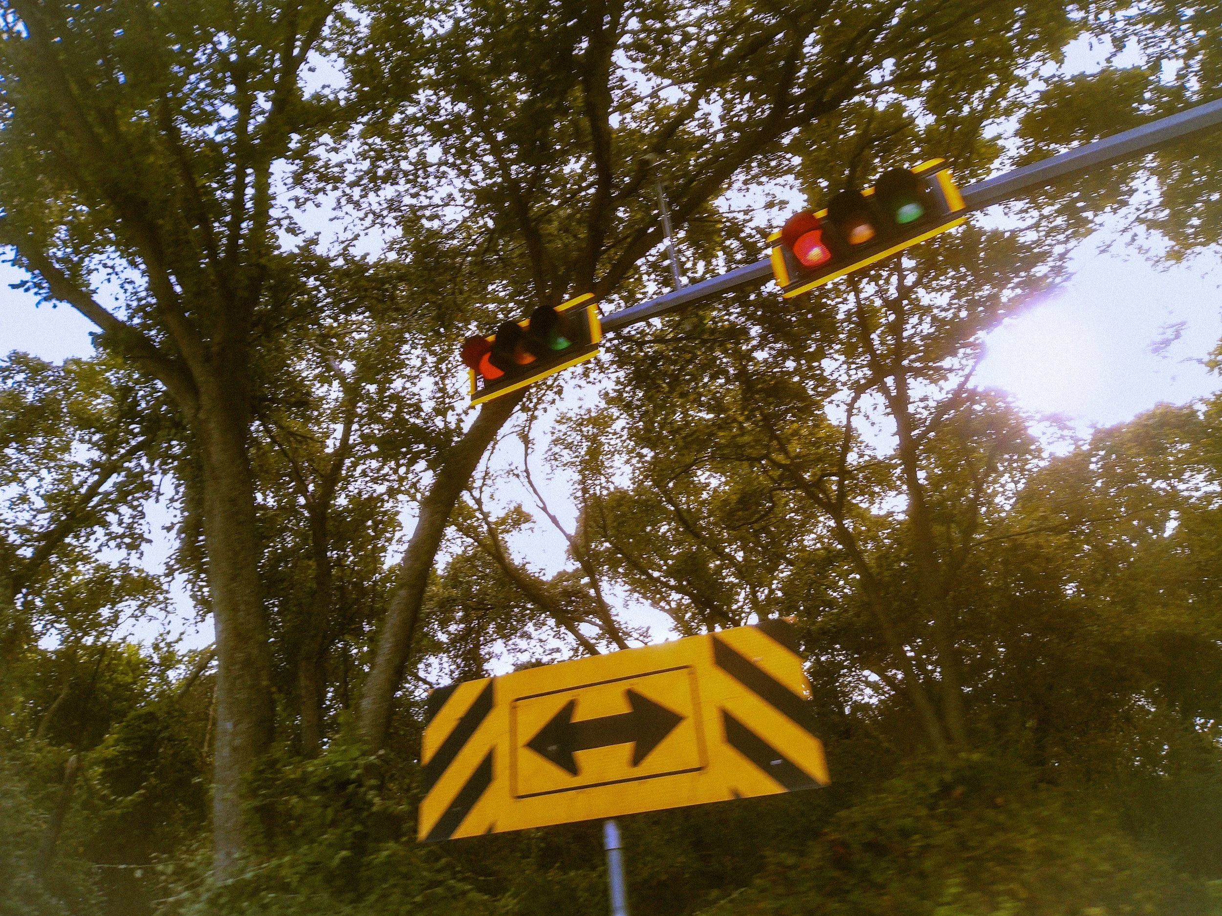 Traffic light hanging over the street showing red, yellow, and green lights with trees in the background and a yellow construction sign with black arrows pointing in different directions.