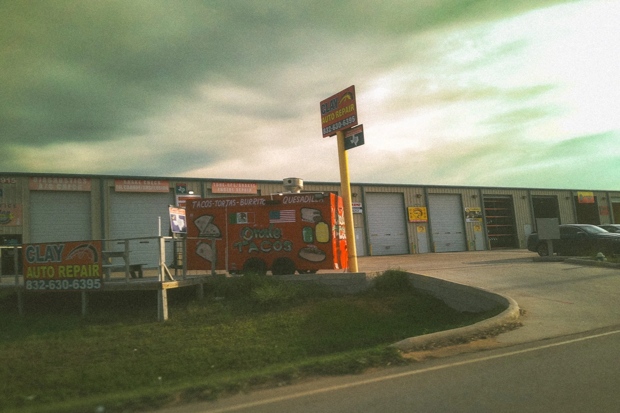 A photo of a commercial building with closed garage doors, a sign for Clay Auto Repair, and a food truck advertising tacos, tortas, burritos, and quesadillas parked in the lot. The sky is cloudy.