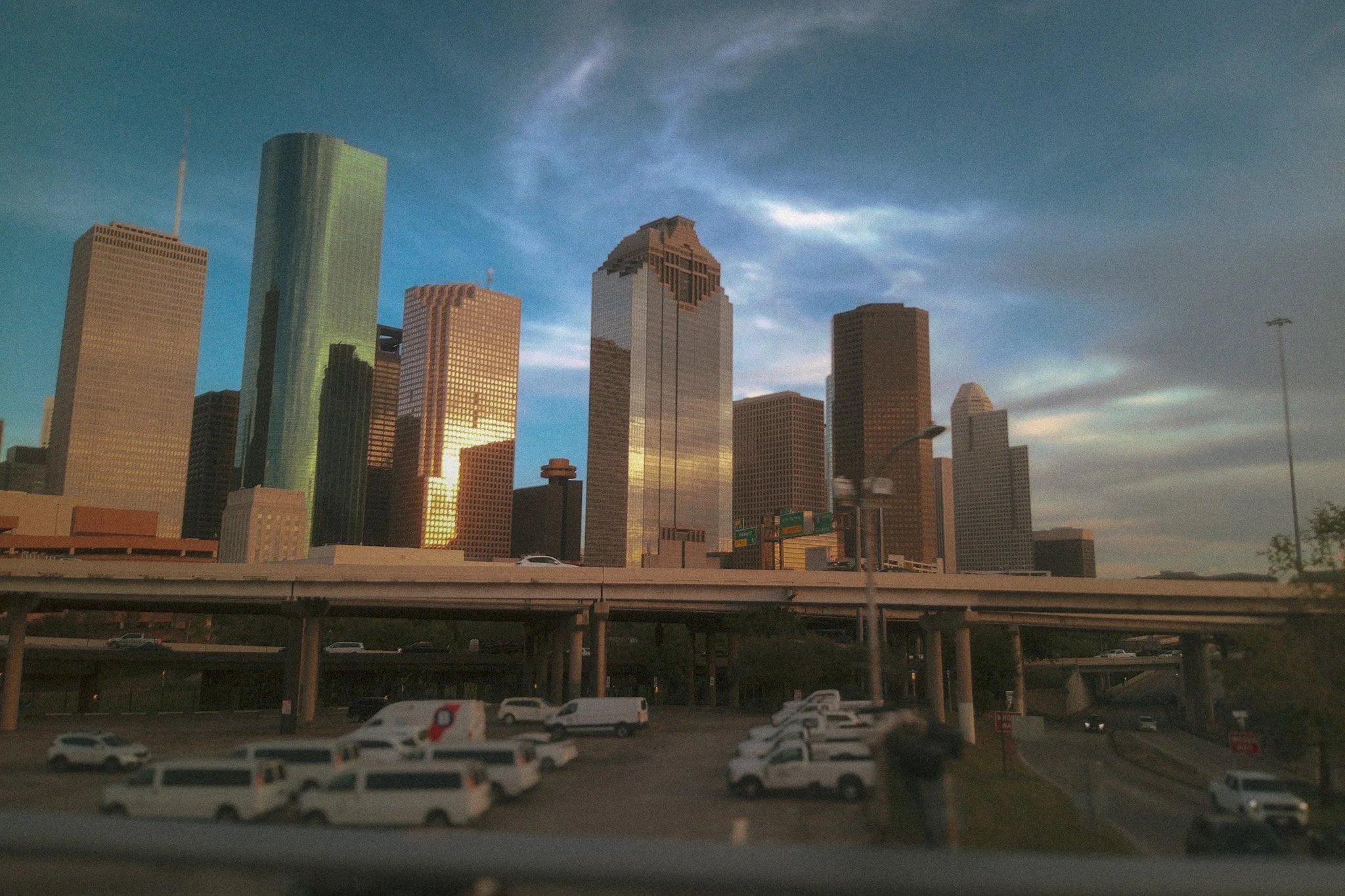 City skyline with tall skyscrapers during sunset, viewed from a highway with parked cars in the foreground.