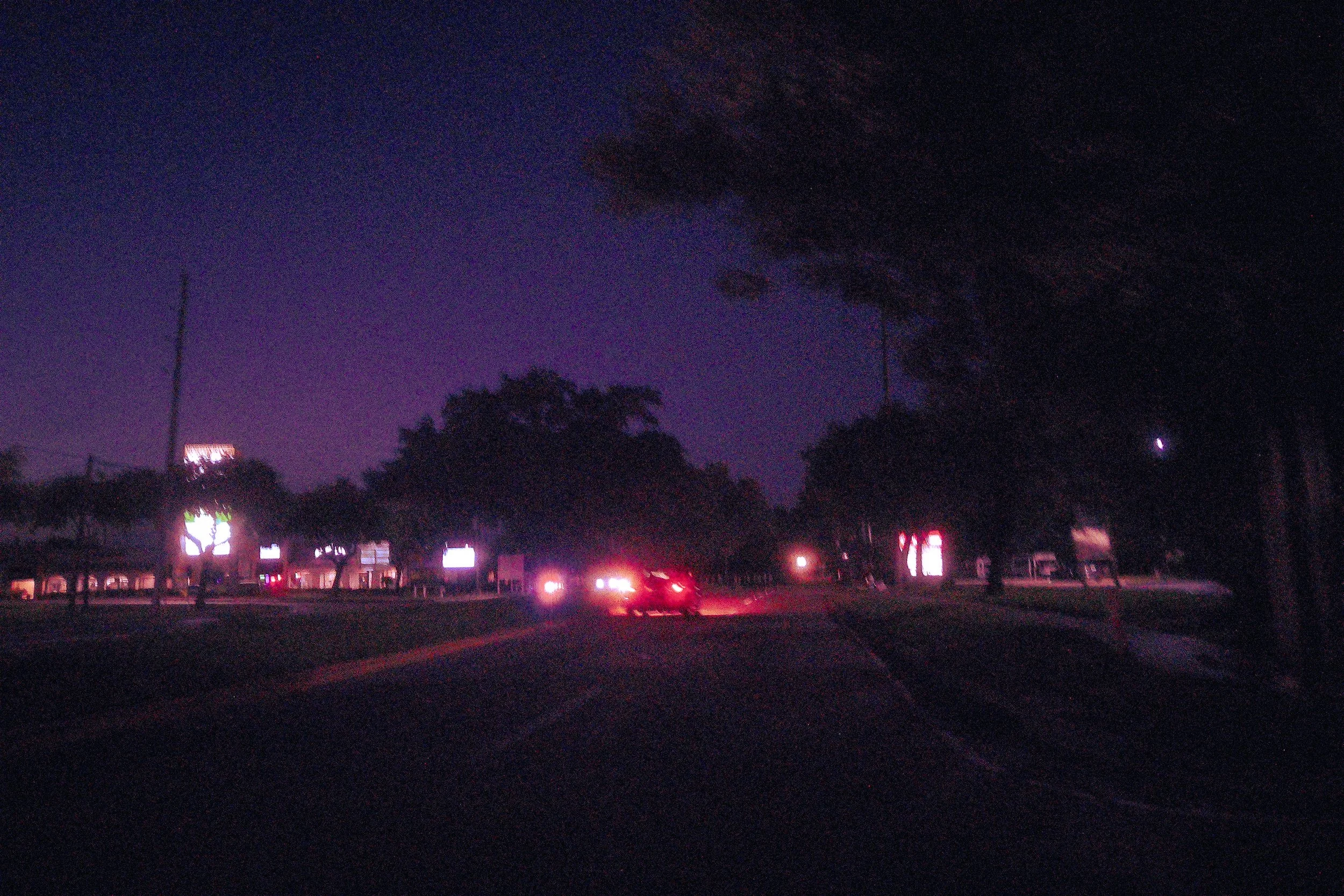 Nighttime scene with dark sky, trees, and illuminated signs, with car headlights and streetlights on a road.