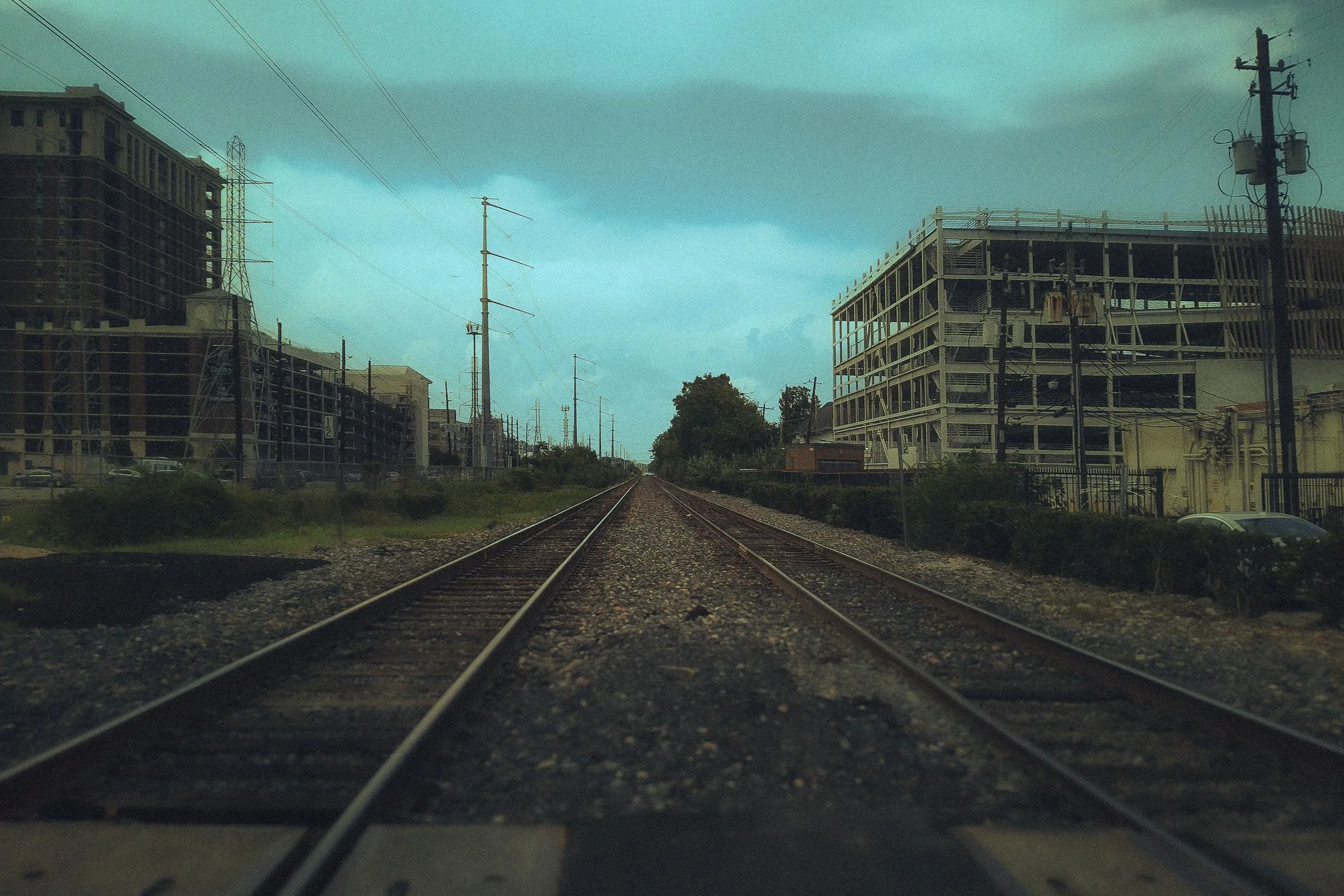 Photo of train tracks leading into the distance with construction sites and buildings on either side, under a cloudy sky.