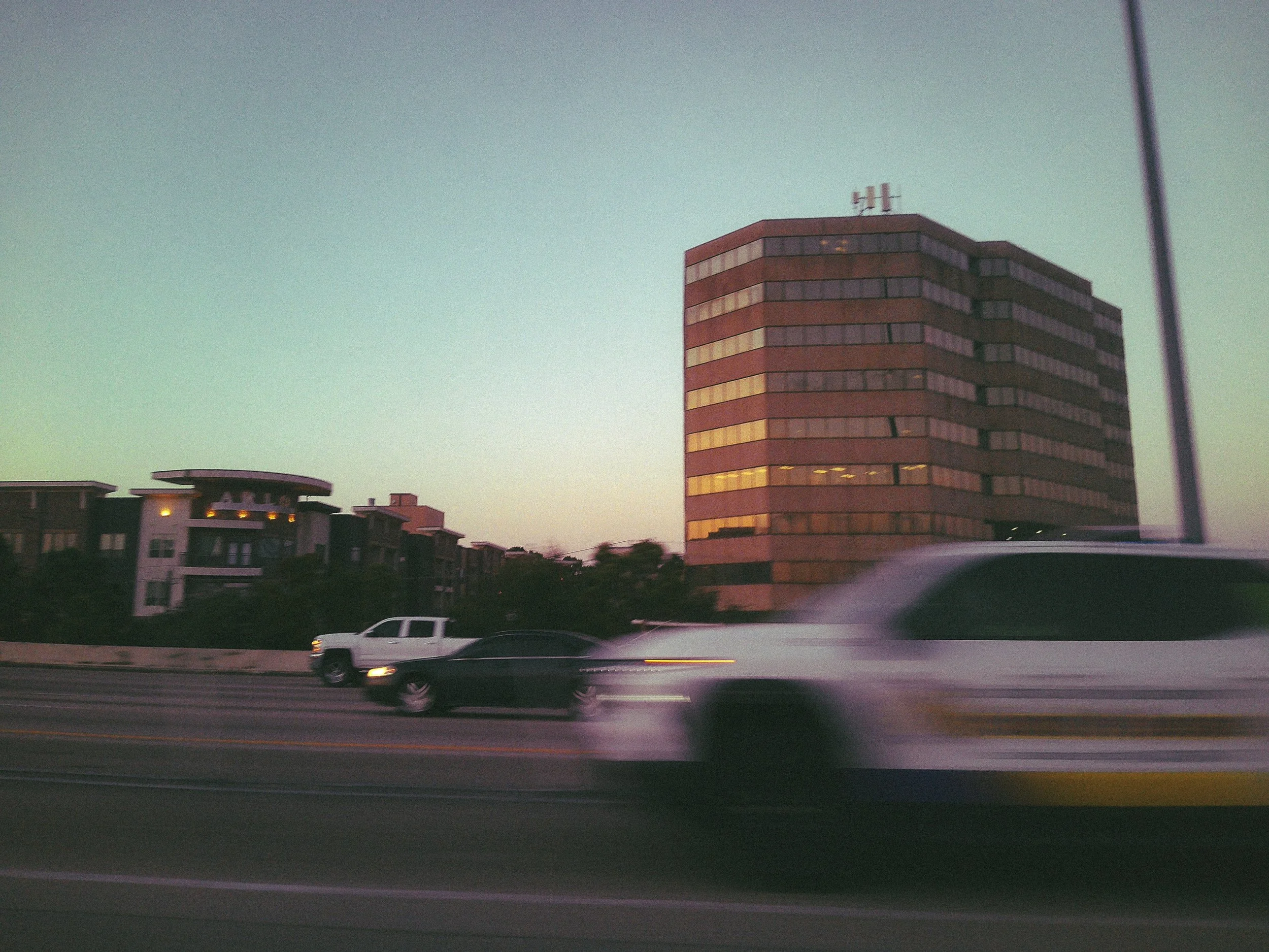 A tall, modern, multi-story building with orange and dark gray horizontal stripes on its exterior, situated next to lower residential buildings. The sky is clear with a gradient from light to darker blue, and there is a street with moving cars in the