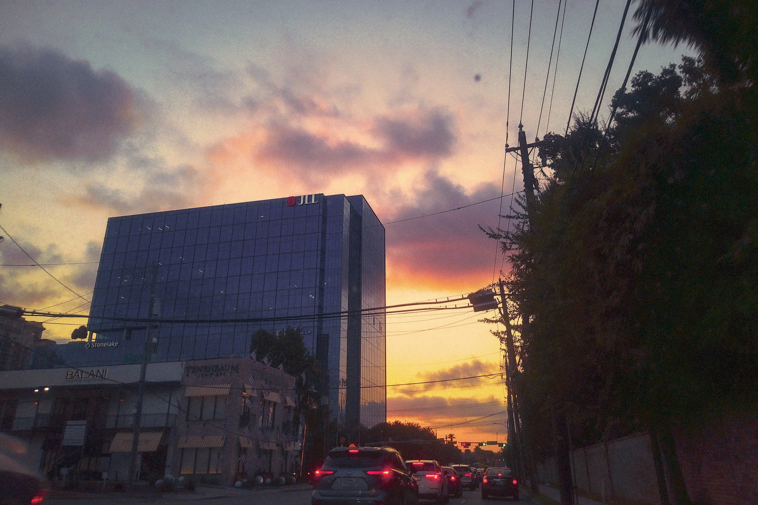 City street at sunrise with cars and a tall glass building with jll logo, power lines, and trees.