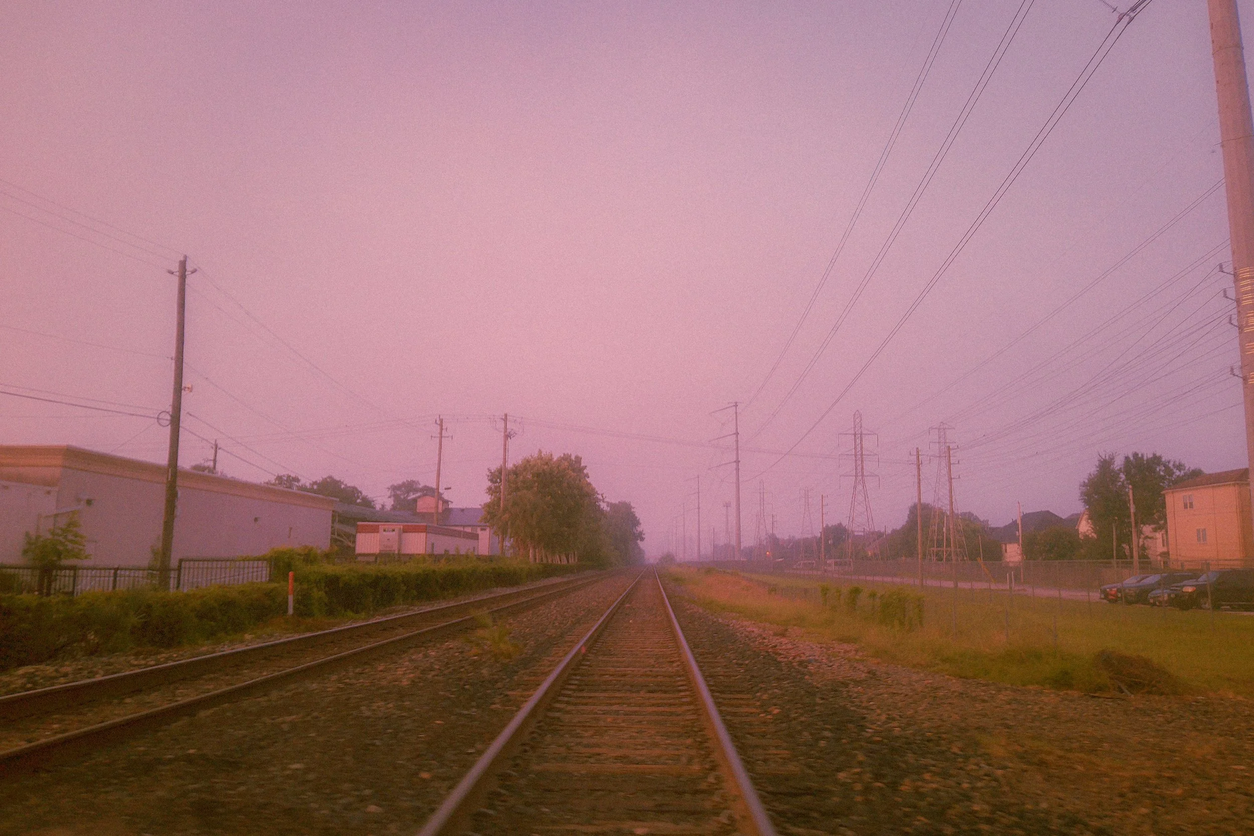 A railway track runs through a suburban area at sunset or sunrise, with power lines and utility poles on either side, and houses and trees in the background under a pinkish sky.