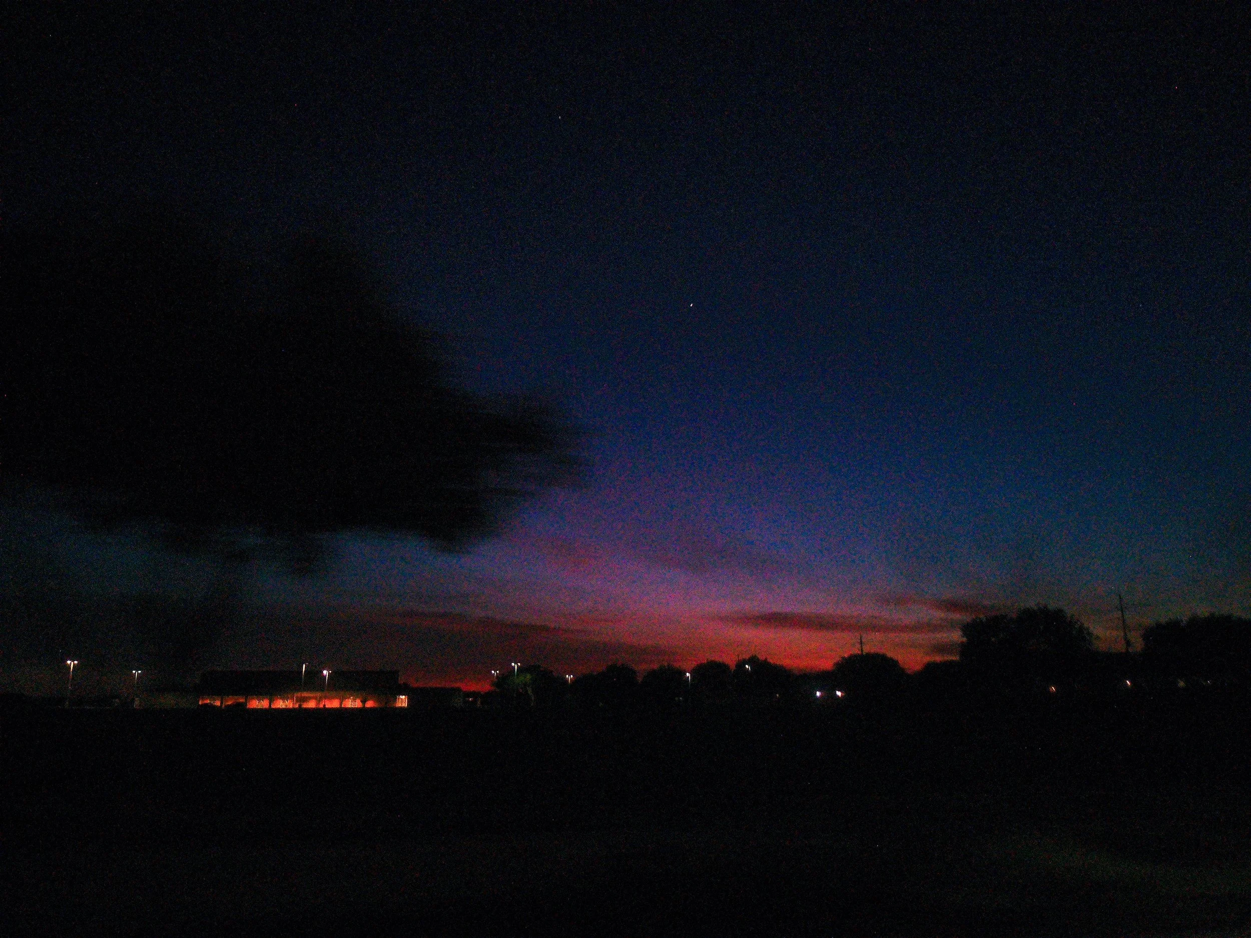 Nighttime sky with faint stars and a colorful horizon with red and purple hues, silhouetted trees and buildings in the foreground.