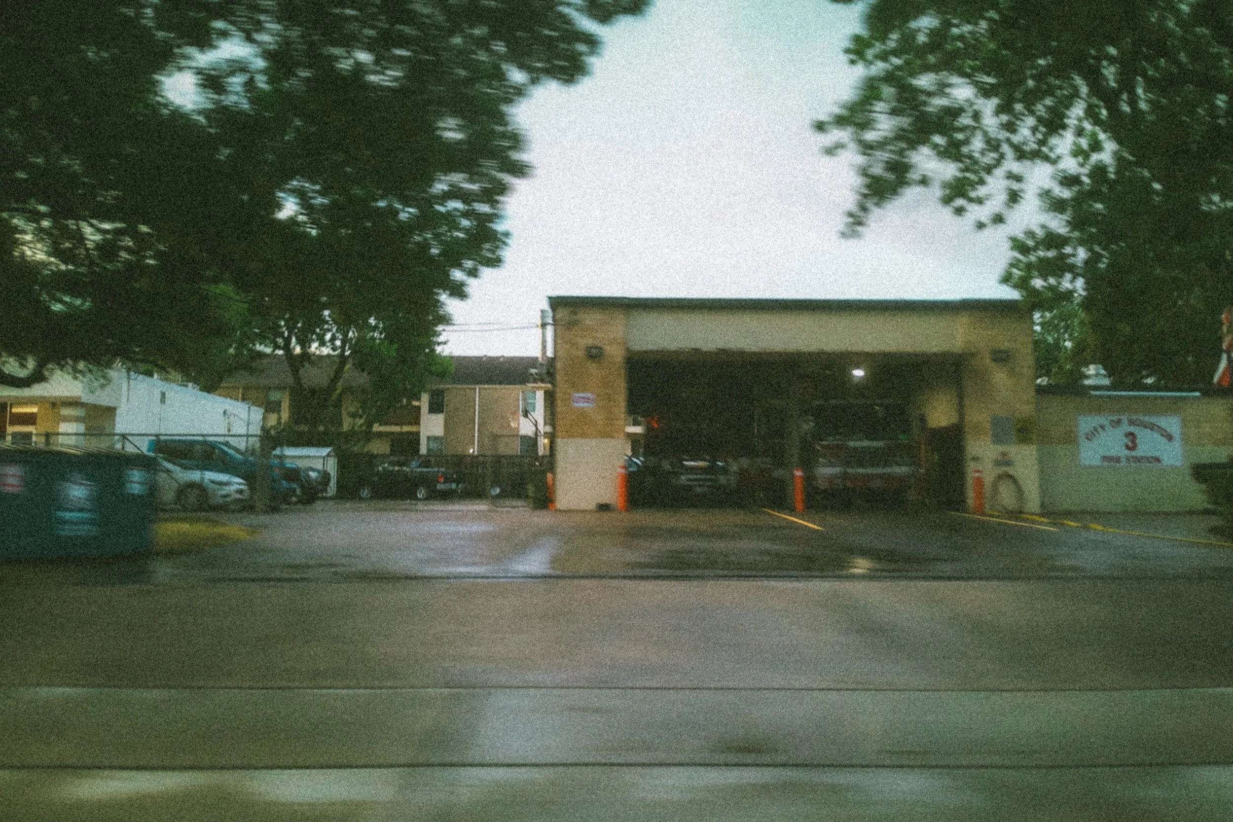 A parking lot with several cars parked in front of a building, large trees surrounding the area, and a partly cloudy sky overhead.