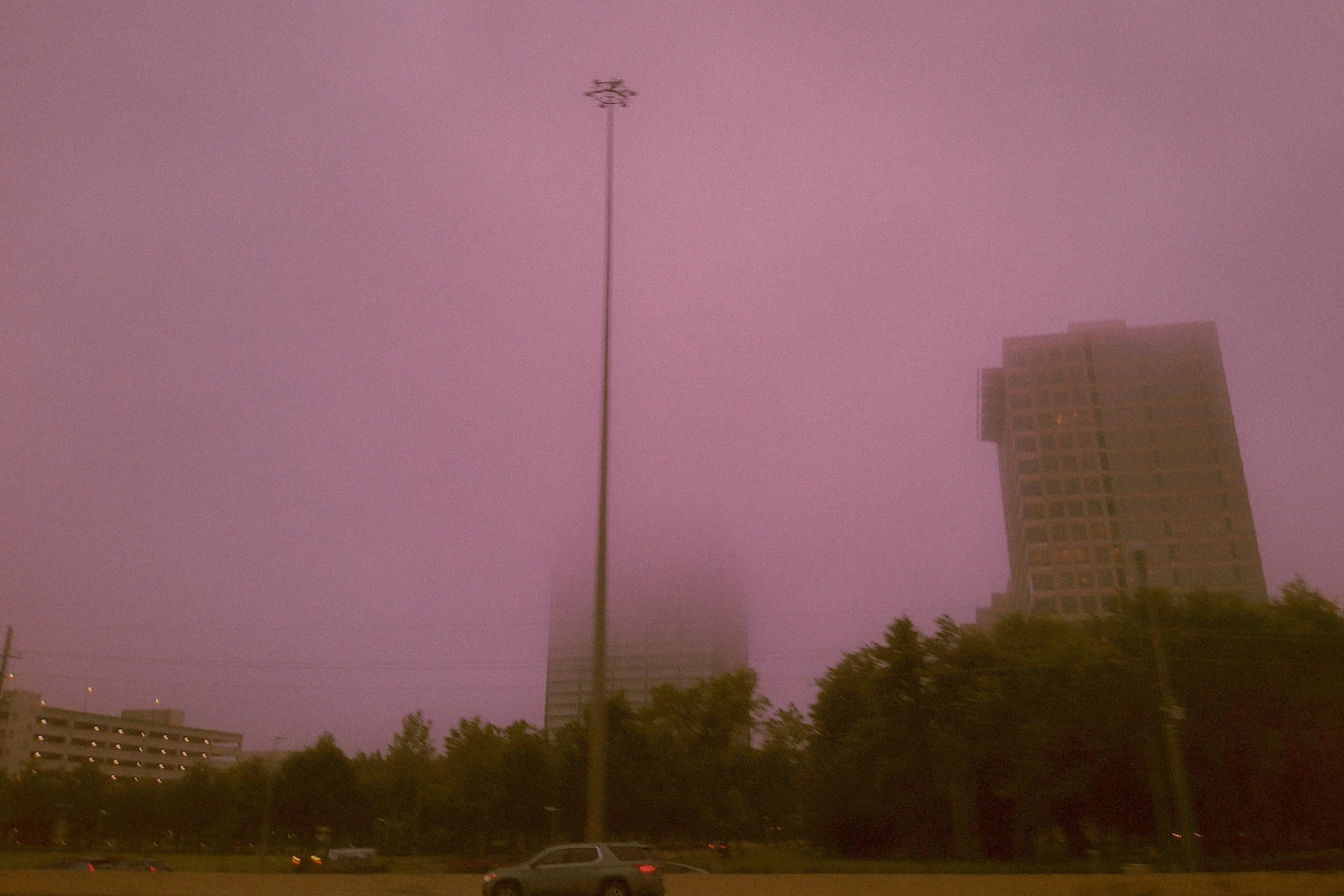 A tall building next to a street lamp against a pinkish sky with some clouds, cars on the road, and trees in the foreground.