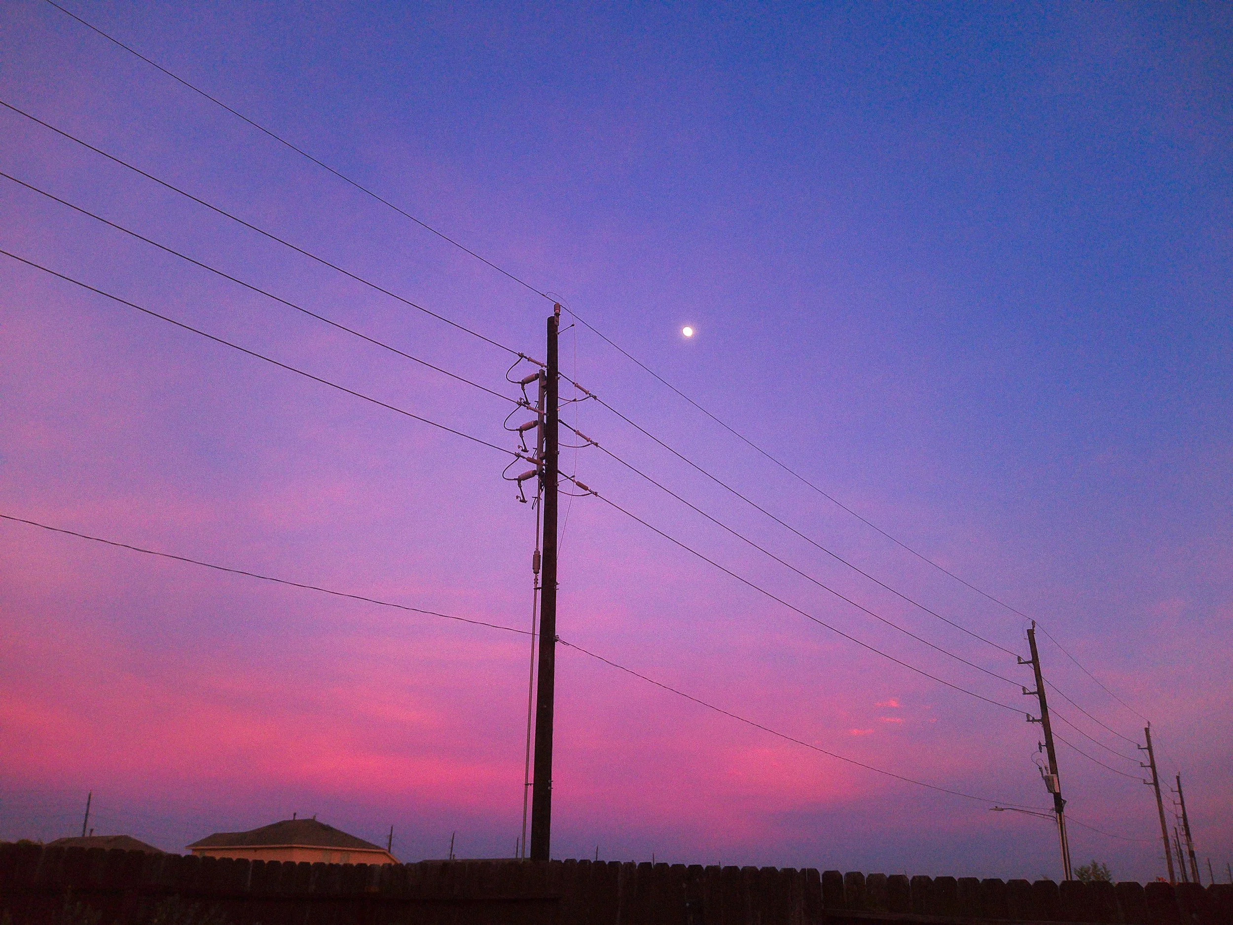 Utility pole with power lines against a twilight sky showing shades of purple, pink, and blue, with a visible moon and silhouettes of houses and trees in the background.