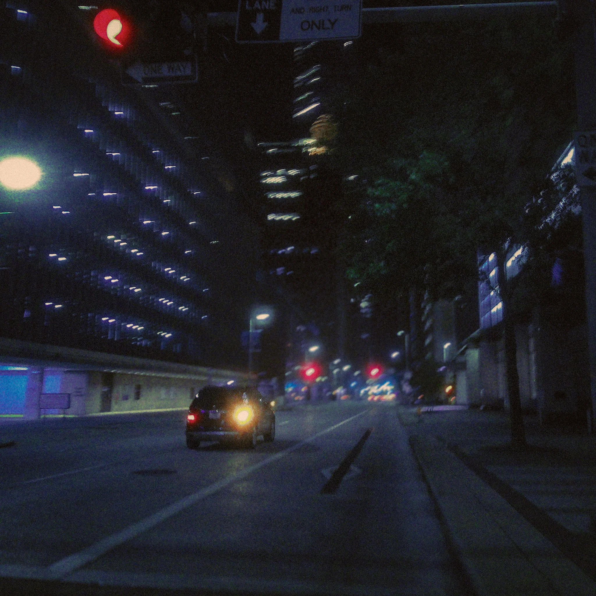 Nighttime view of a street with blurred lights, a vehicle with bright taillights, street signs, and a traffic light in the background.